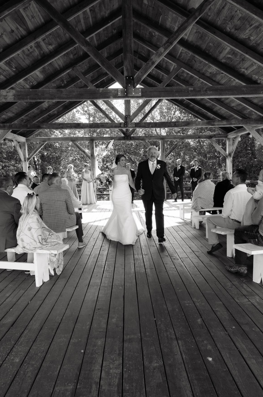 Black and white photo of bride and groom walking down the aisle in a rustic wooden pavilion surrounded by wedding guests.