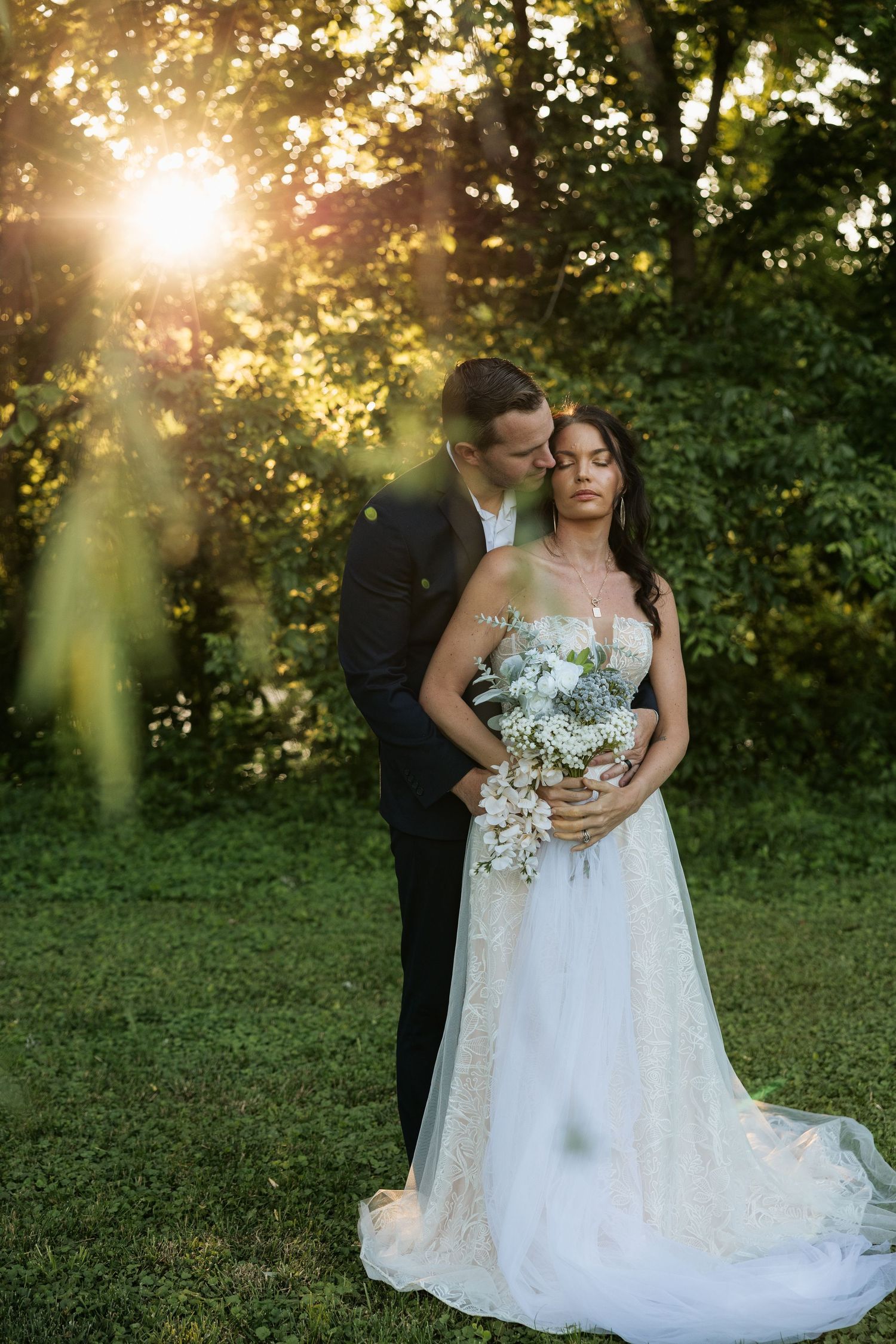 Bride and groom embrace in golden sunset light in lush green garden, bride holding white floral bouquet in elegant wedding gown.