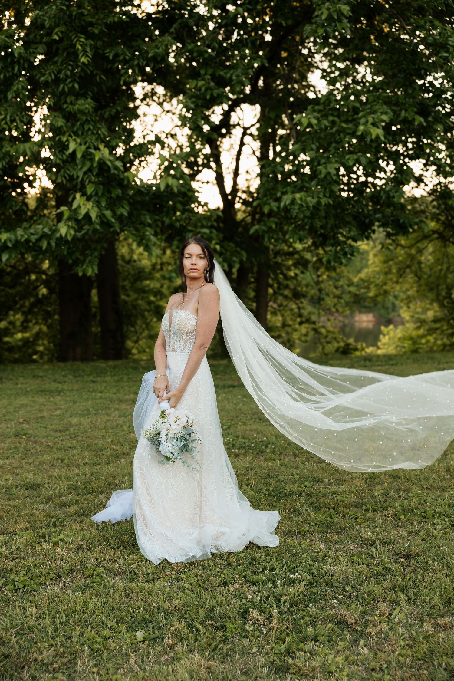 Bride in white lace gown with flowing veil stands on green grass surrounded by trees at golden hour.