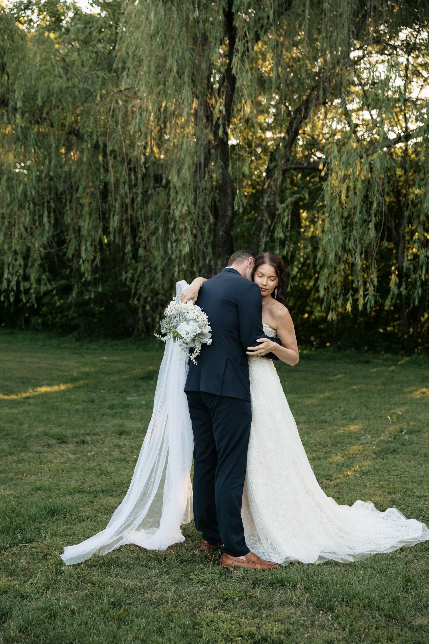 Bride and groom embrace under a weeping willow tree, bride wearing a lace gown with long veil and white bouquet.
