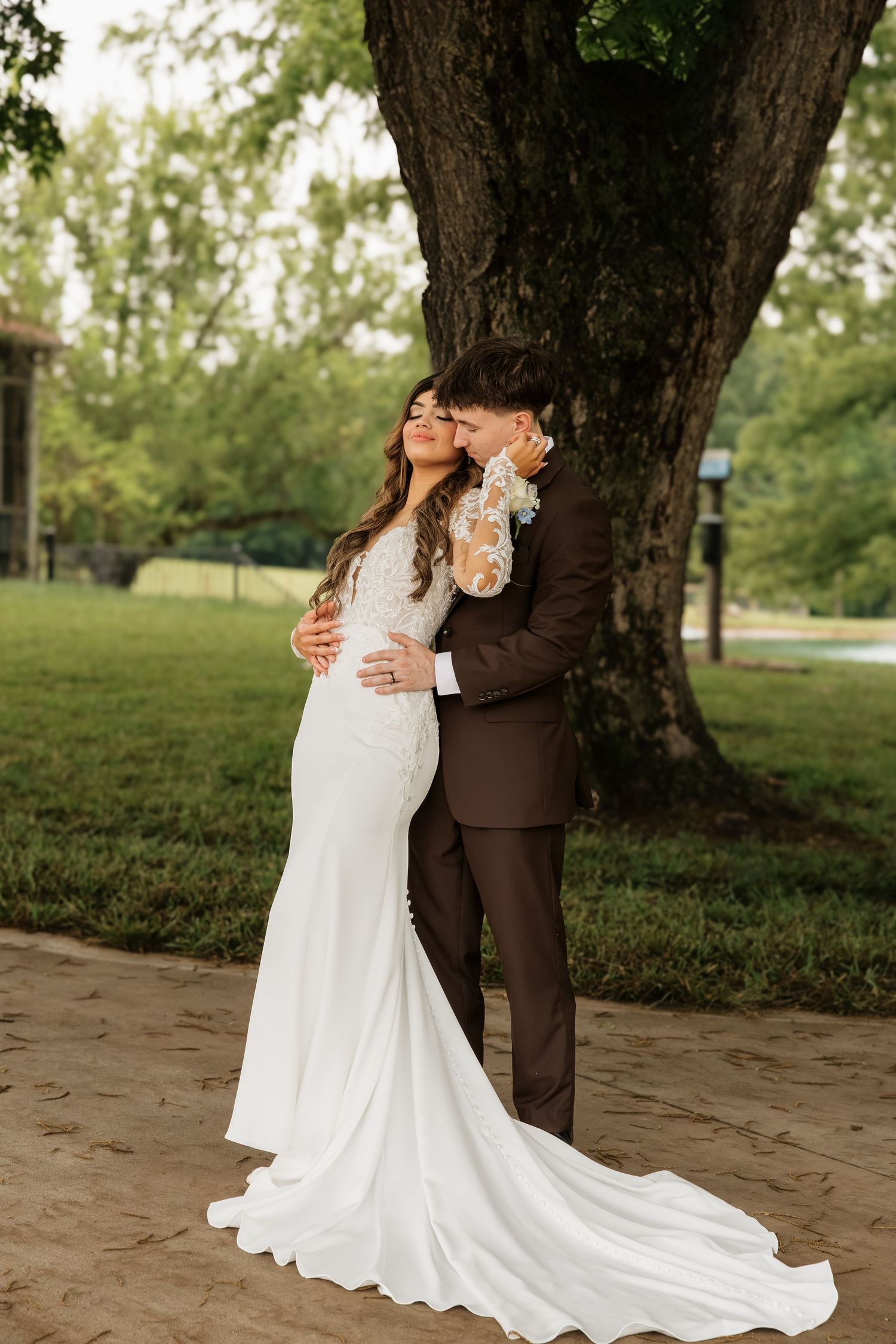 Bride in white lace gown and groom in brown suit embrace under a large tree in a lush green outdoor wedding setting.