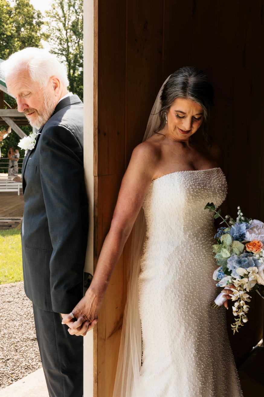 Bride in strapless beaded gown holds bouquet while sharing tender first look moment with older man in suit by barn door.