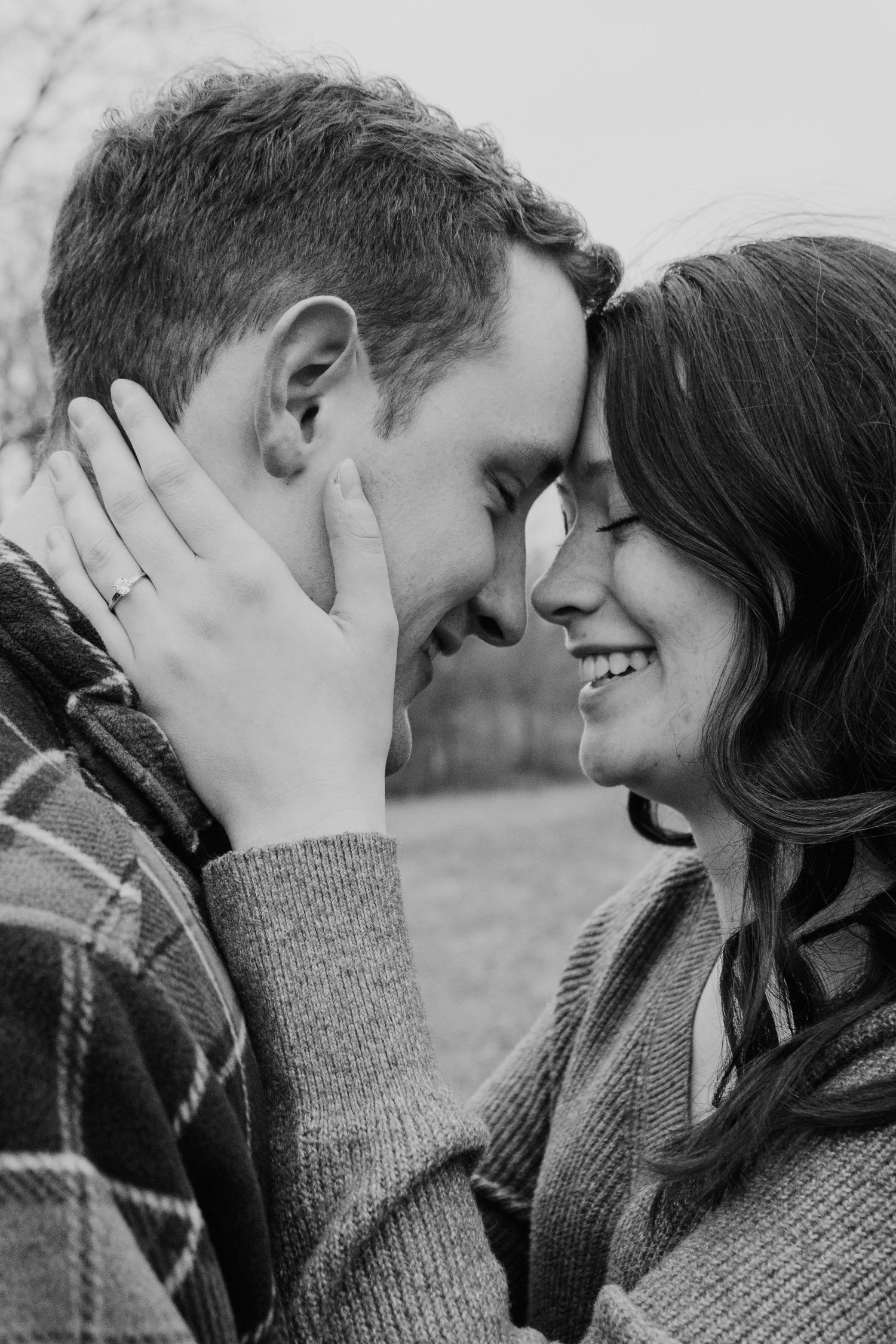 Black and white close-up of smiling couple touching foreheads tenderly outdoors in soft light.