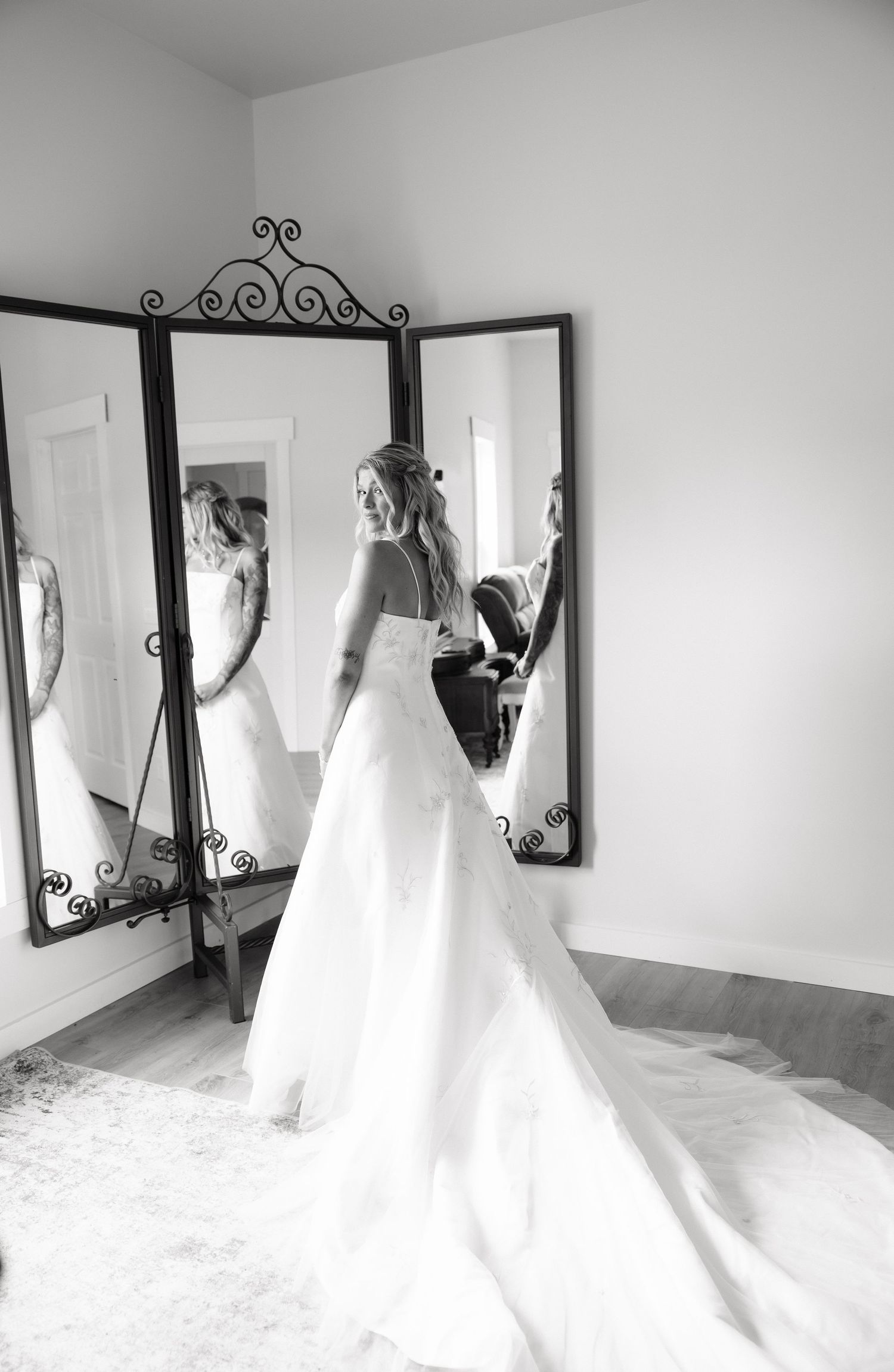 Bride in a flowing white wedding gown stands before a tri-fold mirror in a black and white photo.