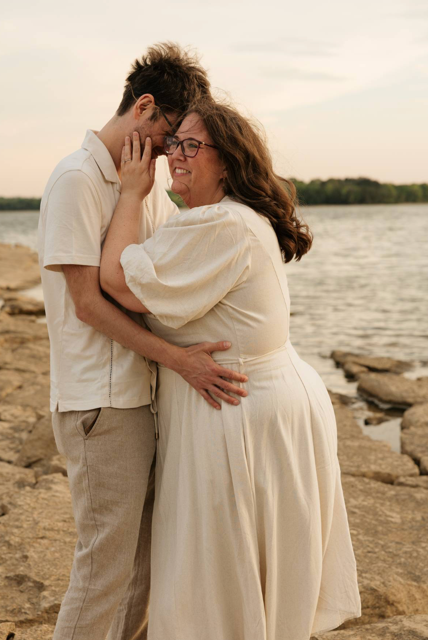 Couple in matching cream outfits embracing romantically by a rocky lakeside at sunset.