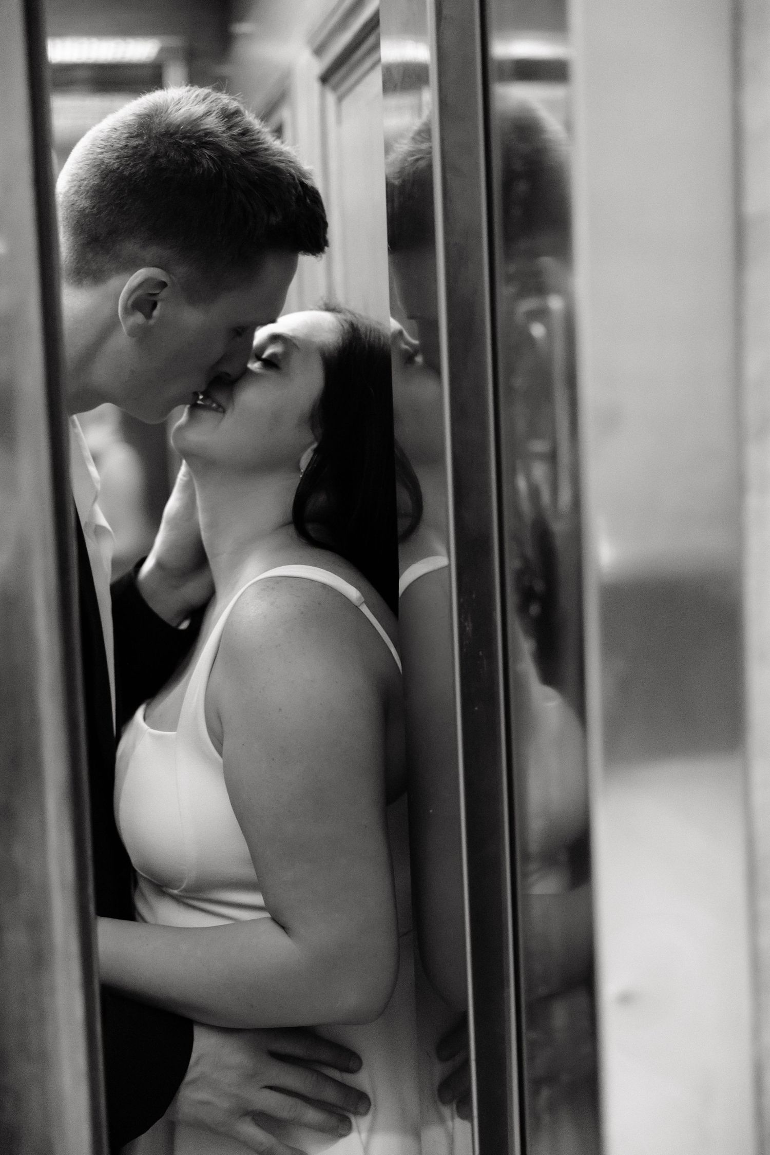 Black and white photo of a couple sharing a romantic kiss near a mirror reflection.