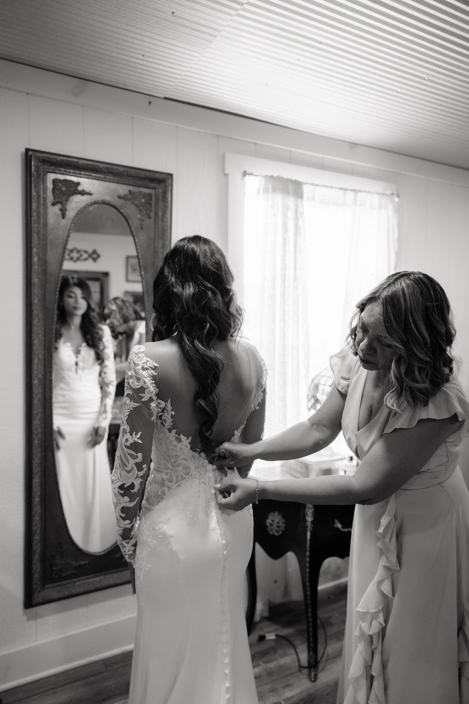 Black and white photo of a bride getting her lace wedding dress fastened by a bridesmaid near a mirror.