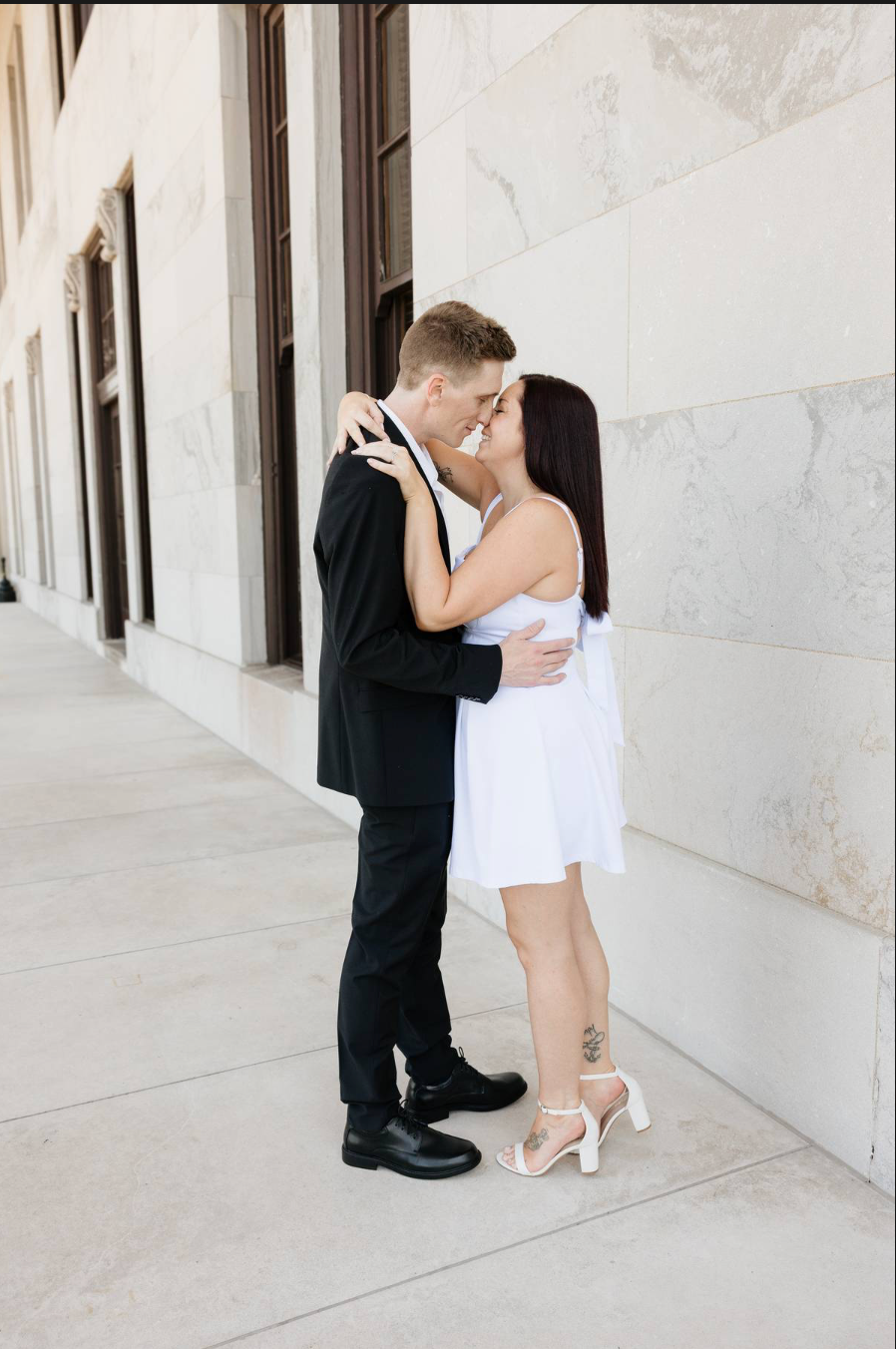 Couple sharing a romantic kiss outdoors, man in black suit and woman in white mini dress with heels against stone wall.