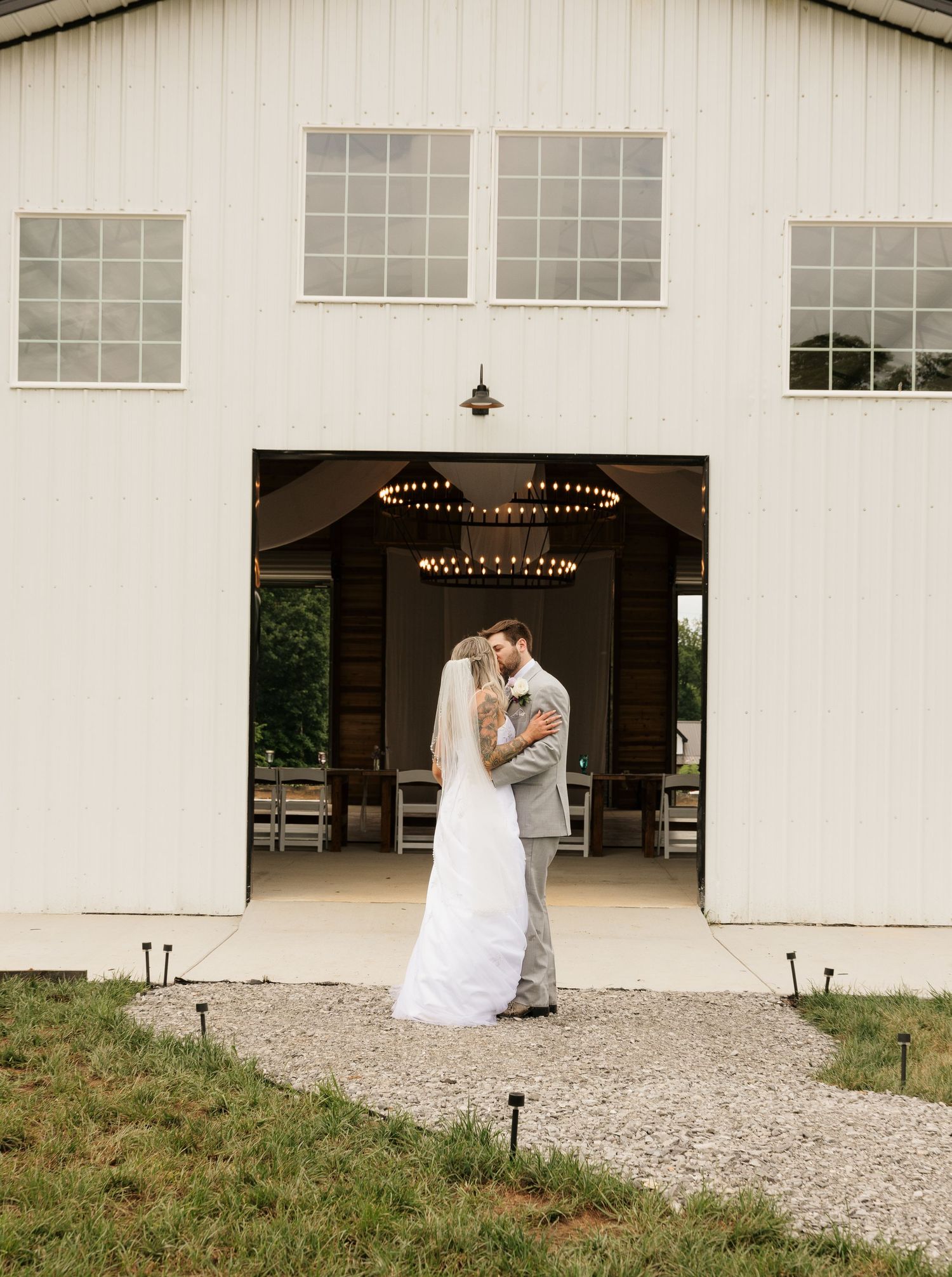 Bride and groom share a romantic embrace outside a white barn wedding venue with elegant chandeliers and string lights.