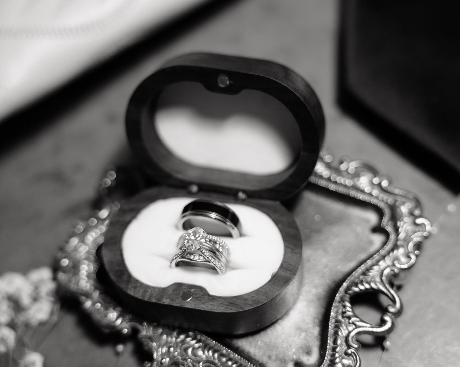 Black and white photo of diamond wedding rings displayed in a round jewelry box with chain necklace nearby.