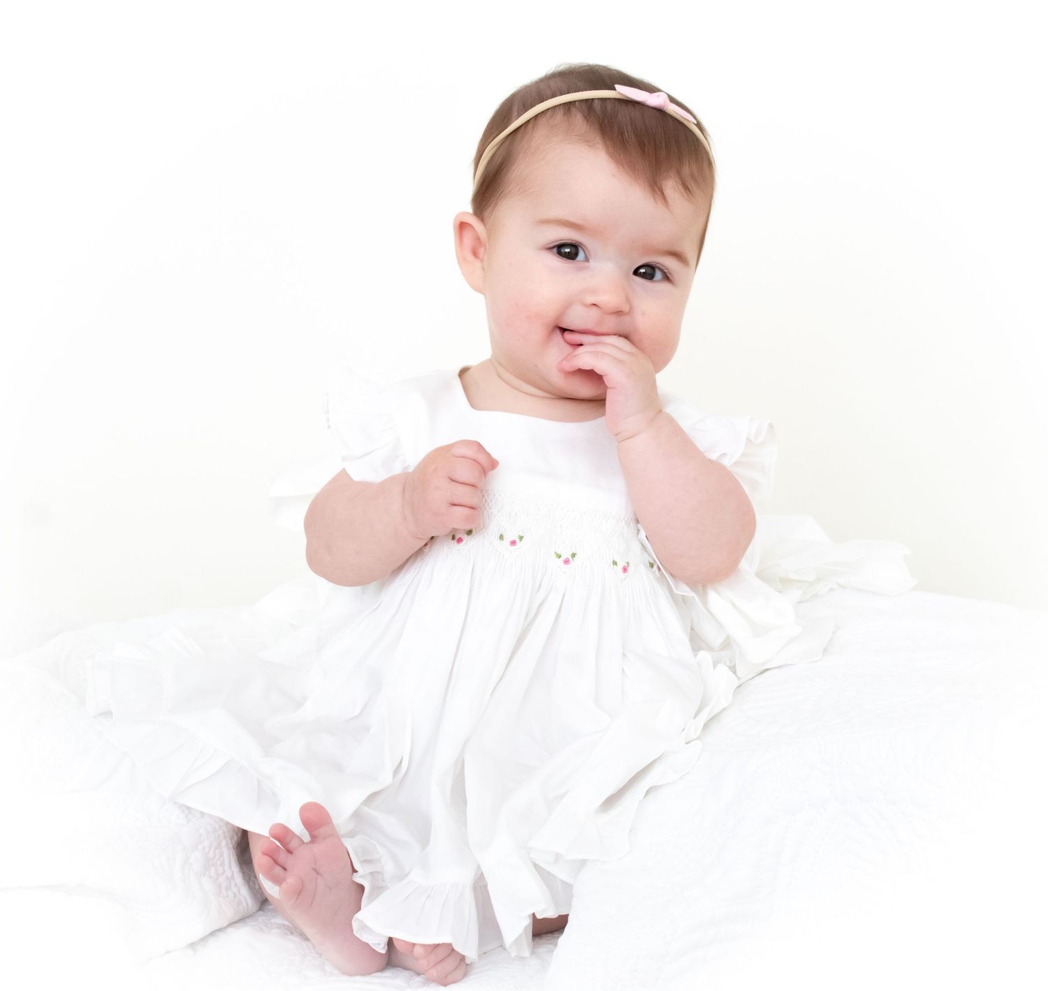 Adorable baby girl in white dress sitting on white blanket, finger in mouth, smiling.