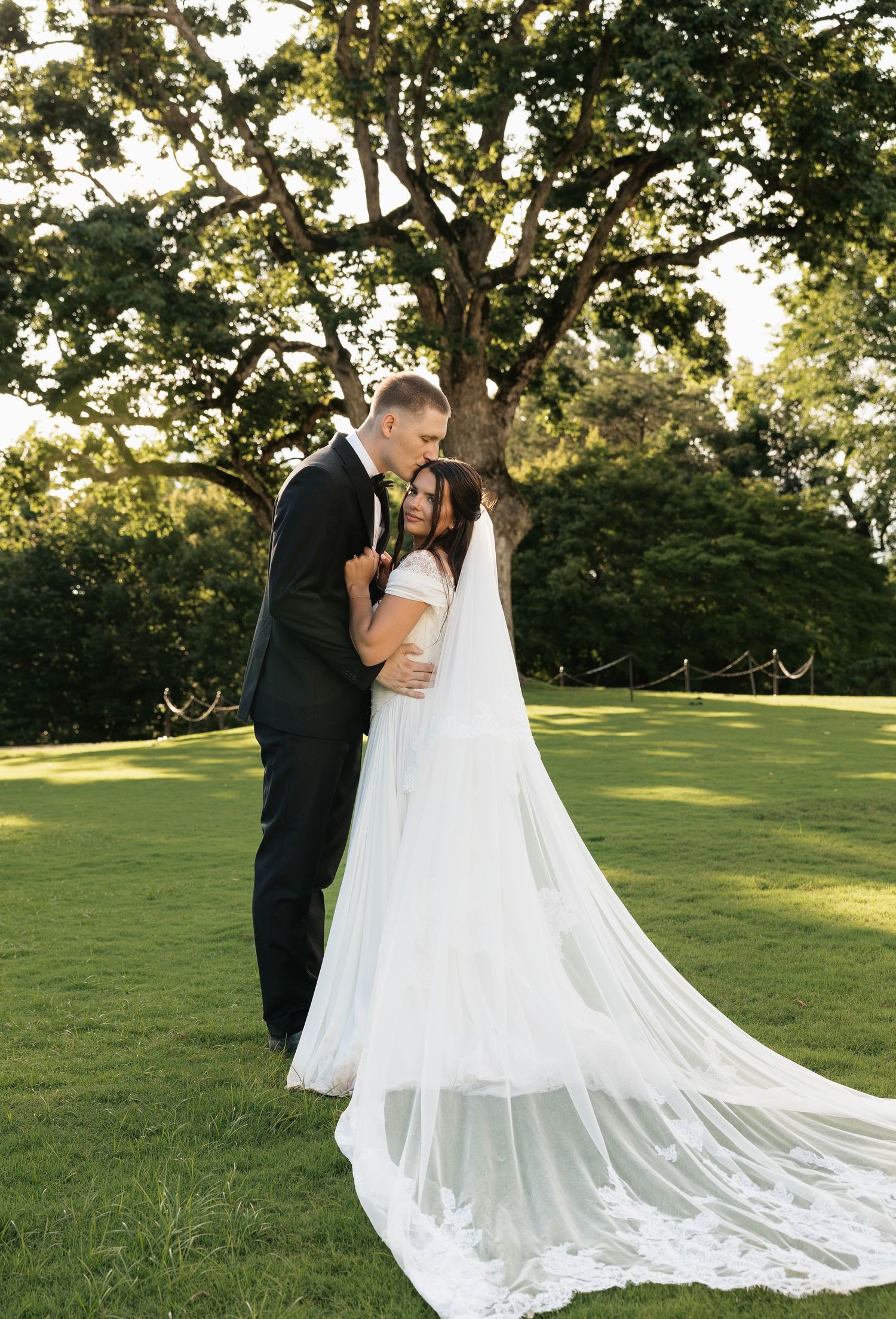Bride and groom share a romantic embrace on green lawn, with bride in flowing white gown and cathedral veil outdoors.