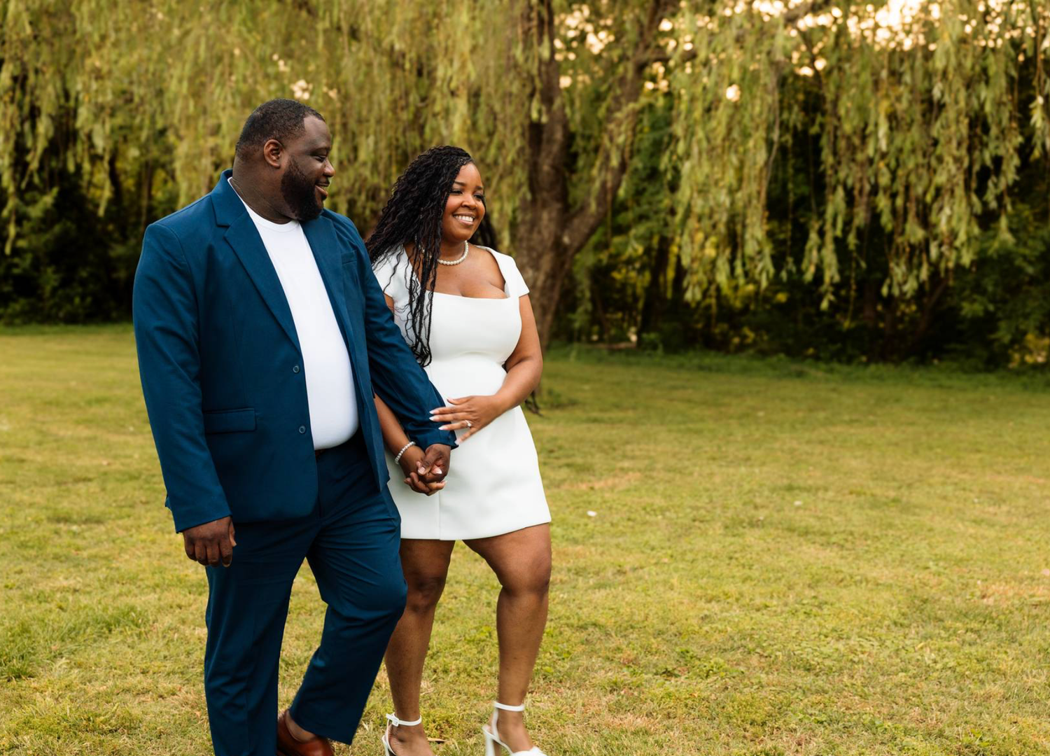 A smiling Black couple walks hand-in-hand outdoors, man in blue suit, woman in white dress, with lush green trees behind.