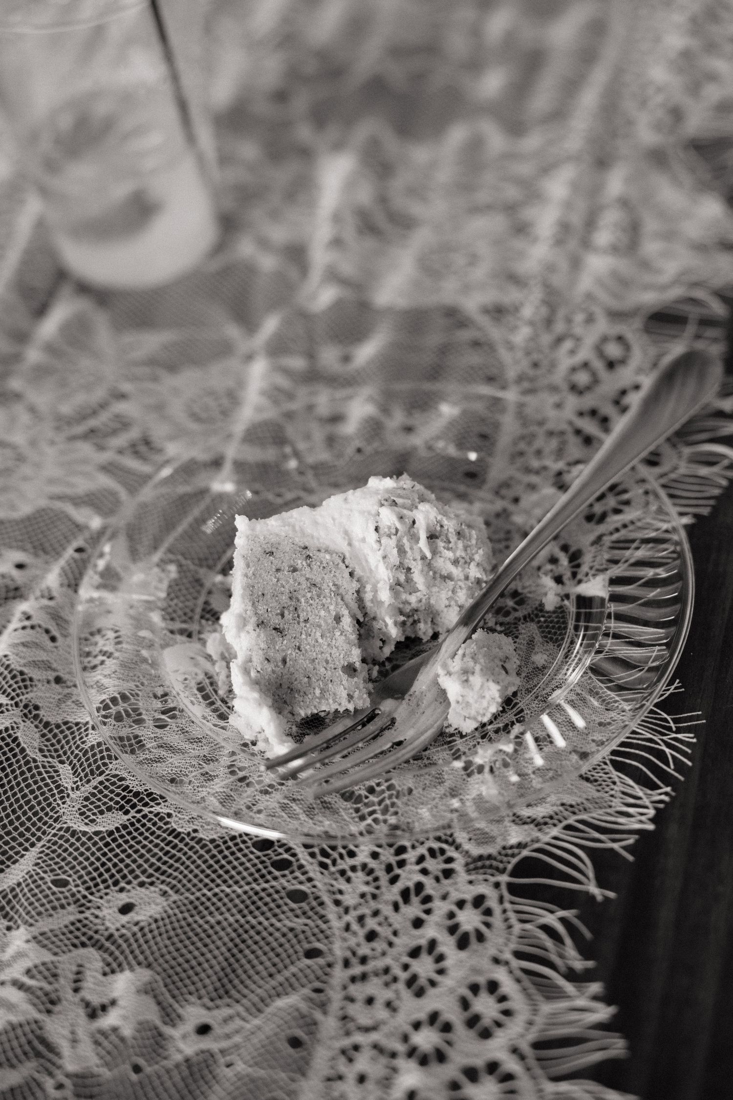 Black and white photo of a slice of cake on a clear glass plate with a fork, on a lace tablecloth.