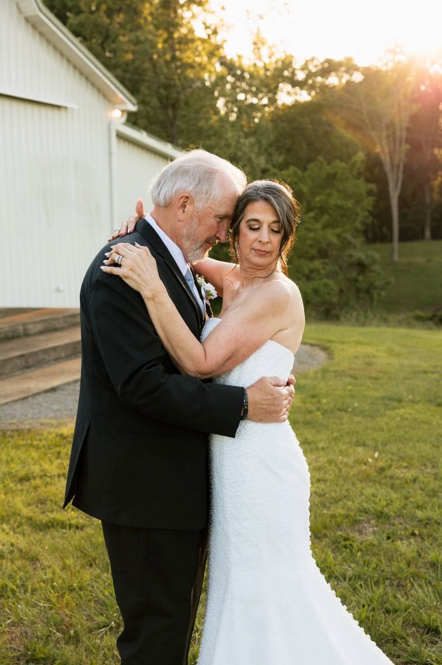 Bride in white strapless gown embraces older man in black tuxedo during golden hour outdoor wedding portrait.