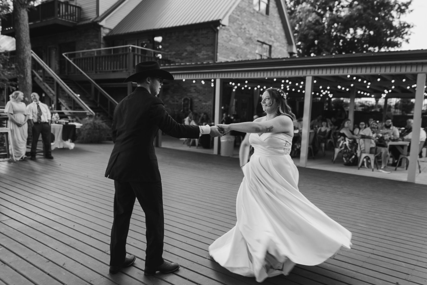 Groom in cowboy hat twirls bride in flowing white dress on outdoor dance floor at wedding reception.