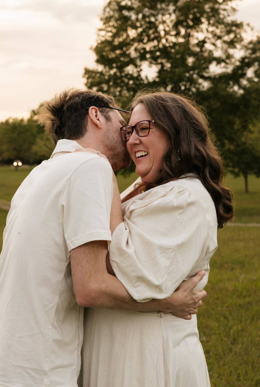 A laughing woman in a white dress embraces a man whispering in her ear outdoors at golden hour.