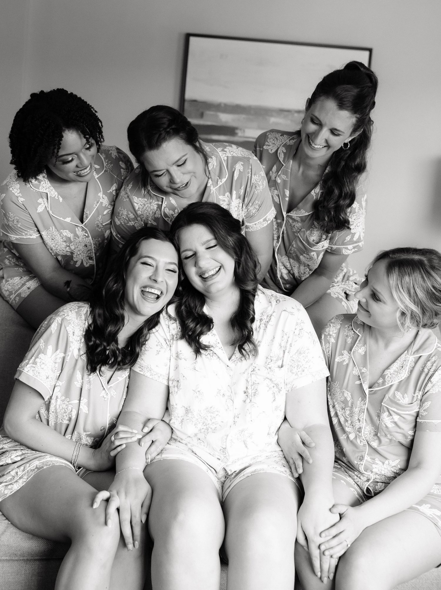 Black and white photo of five women in matching robes laughing together on a couch during bridal party getting ready.