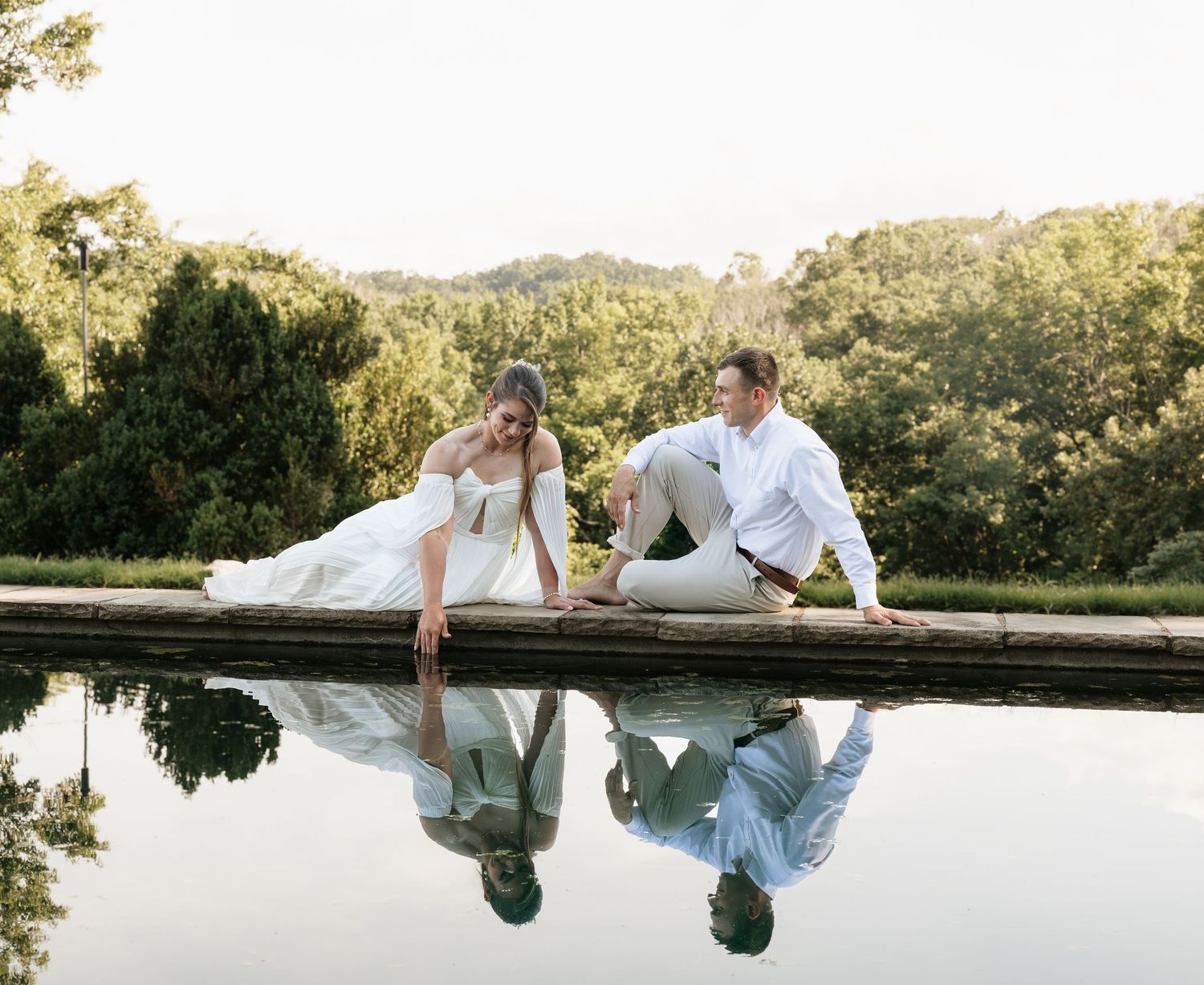 Bride and groom sitting by a reflective pool surrounded by lush greenery on their wedding day.