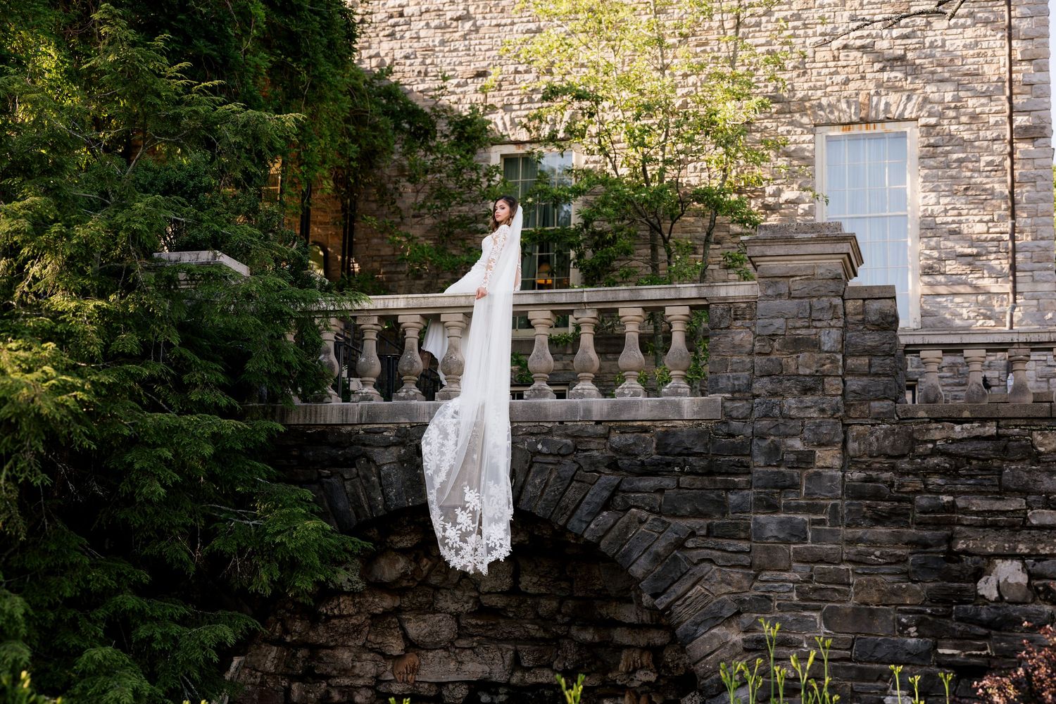 Bride in white wedding gown with long lace veil stands on stone balcony bridge surrounded by lush greenery.