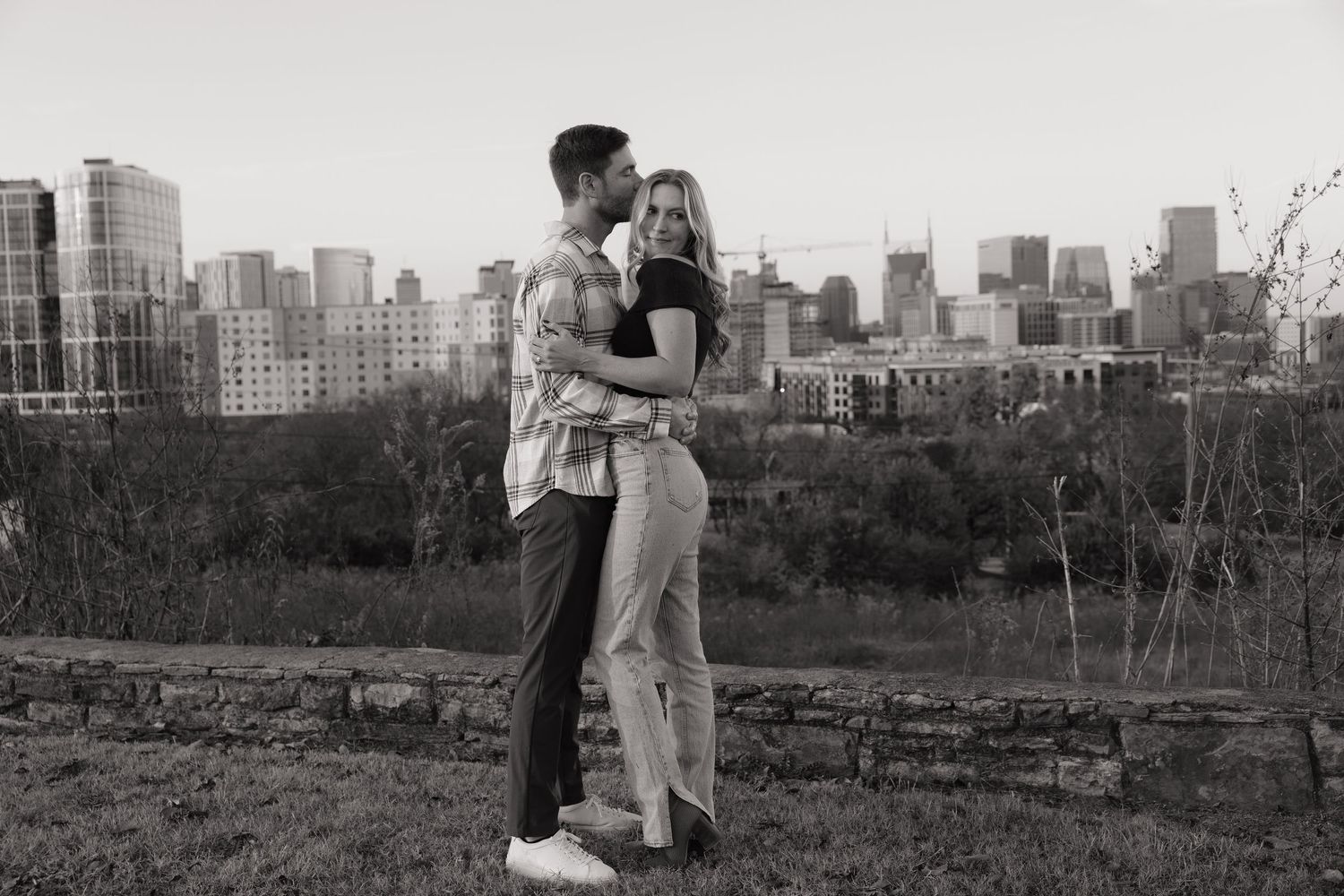 Black and white photo of couple embracing with Nashville city skyline visible in the background at dusk.