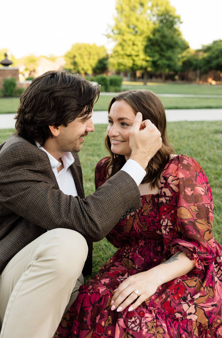 A couple shares a tender moment on grass, man gently touching womans face, woman in floral red dress smiling warmly.