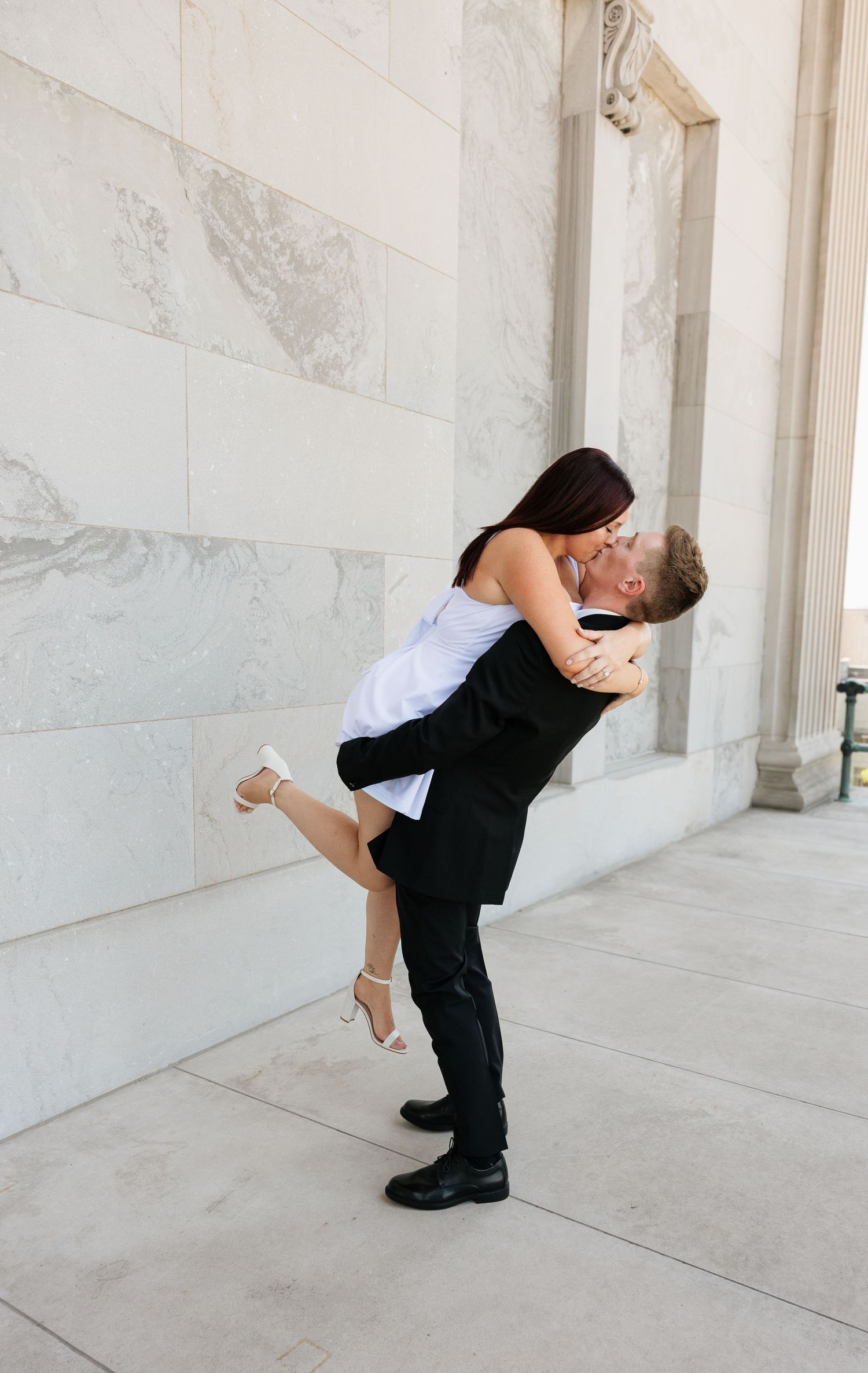 A groom lifts his bride in a romantic kiss outside a marble building in a classic wedding portrait.