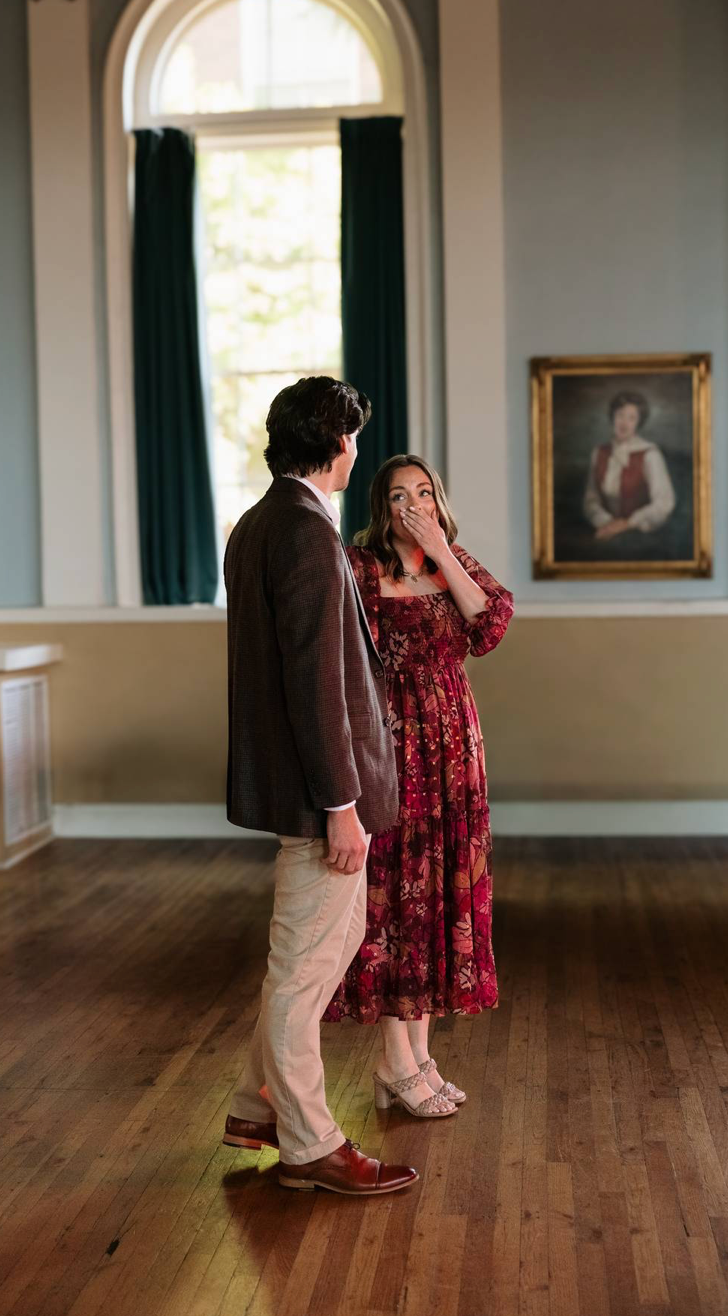 Man surprising woman in red floral dress during proposal in elegant hall.