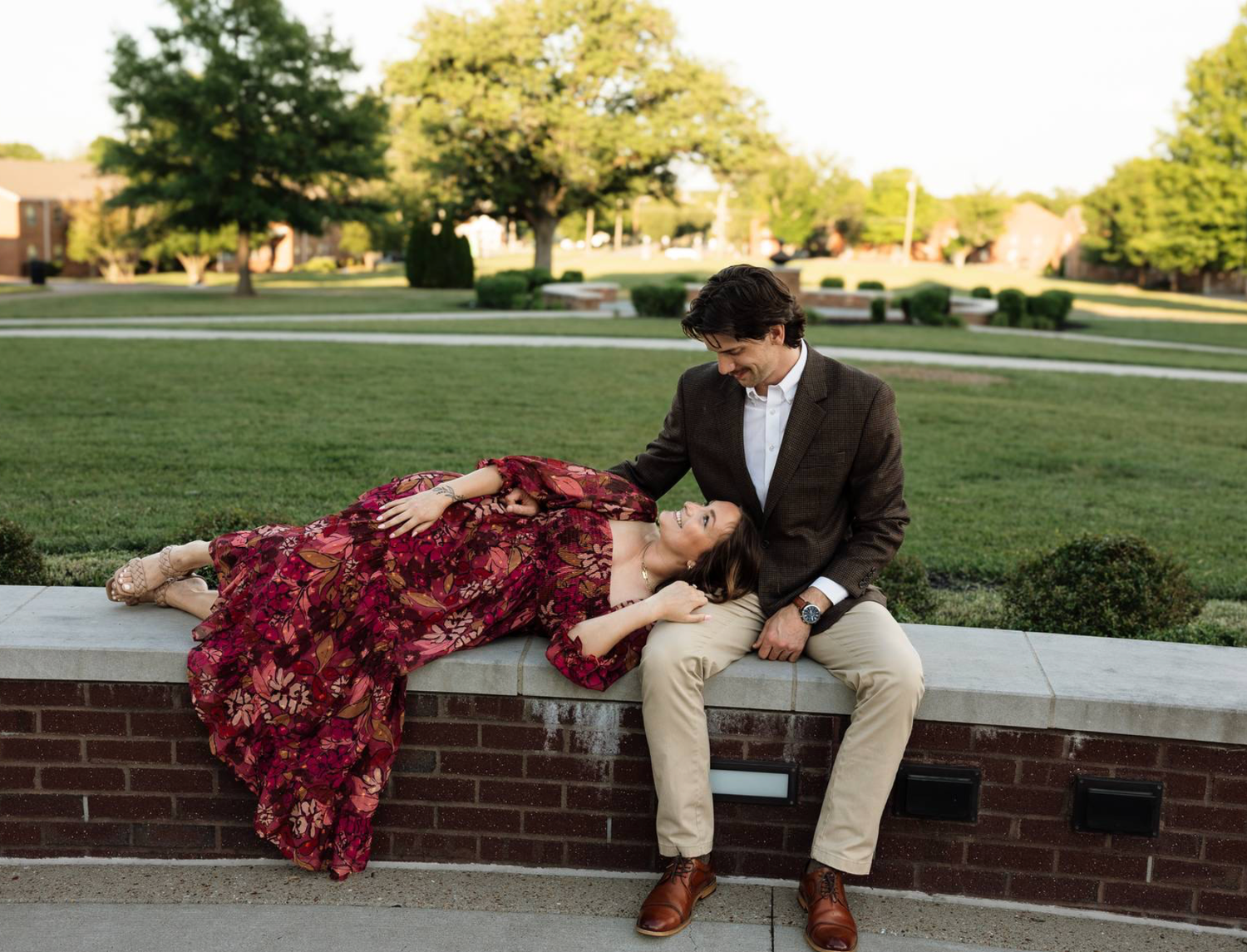 Couple posing on brick wall, woman in floral red dress lying down, man in brown blazer, green park setting.