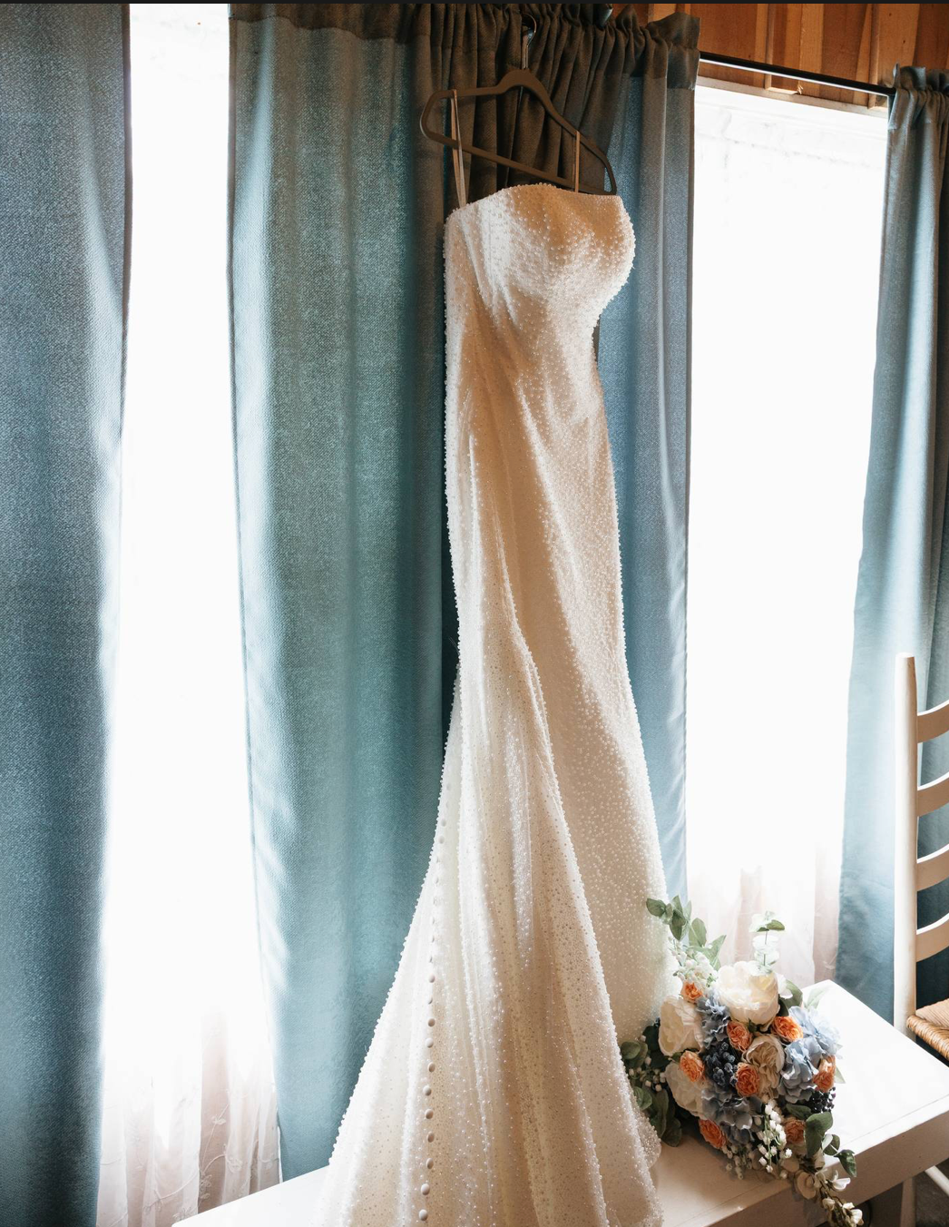 Elegant white lace wedding gown hanging on a wooden hanger between blue curtains with a floral bouquet below.