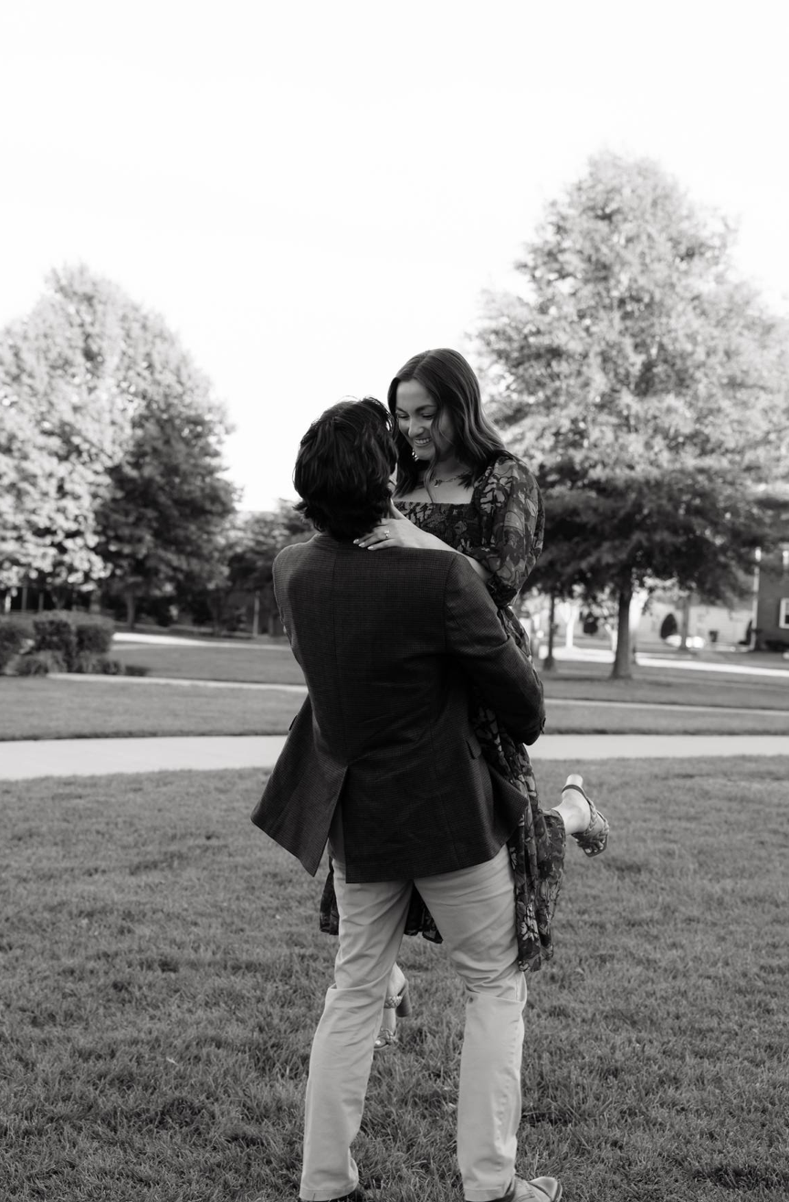Black and white photo of a couple embracing outdoors, man lifting woman in a romantic moment on a green lawn.