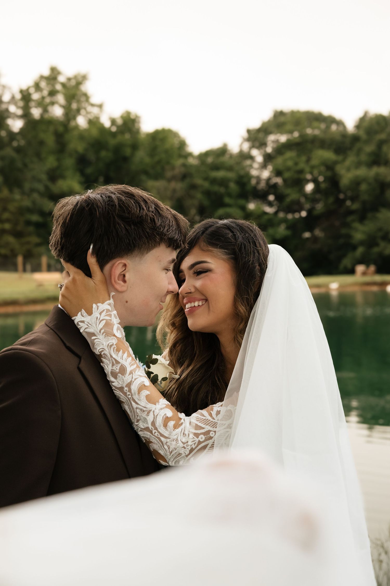 A smiling bride in lace gown and veil touches foreheads with groom in dark suit near a serene lake.