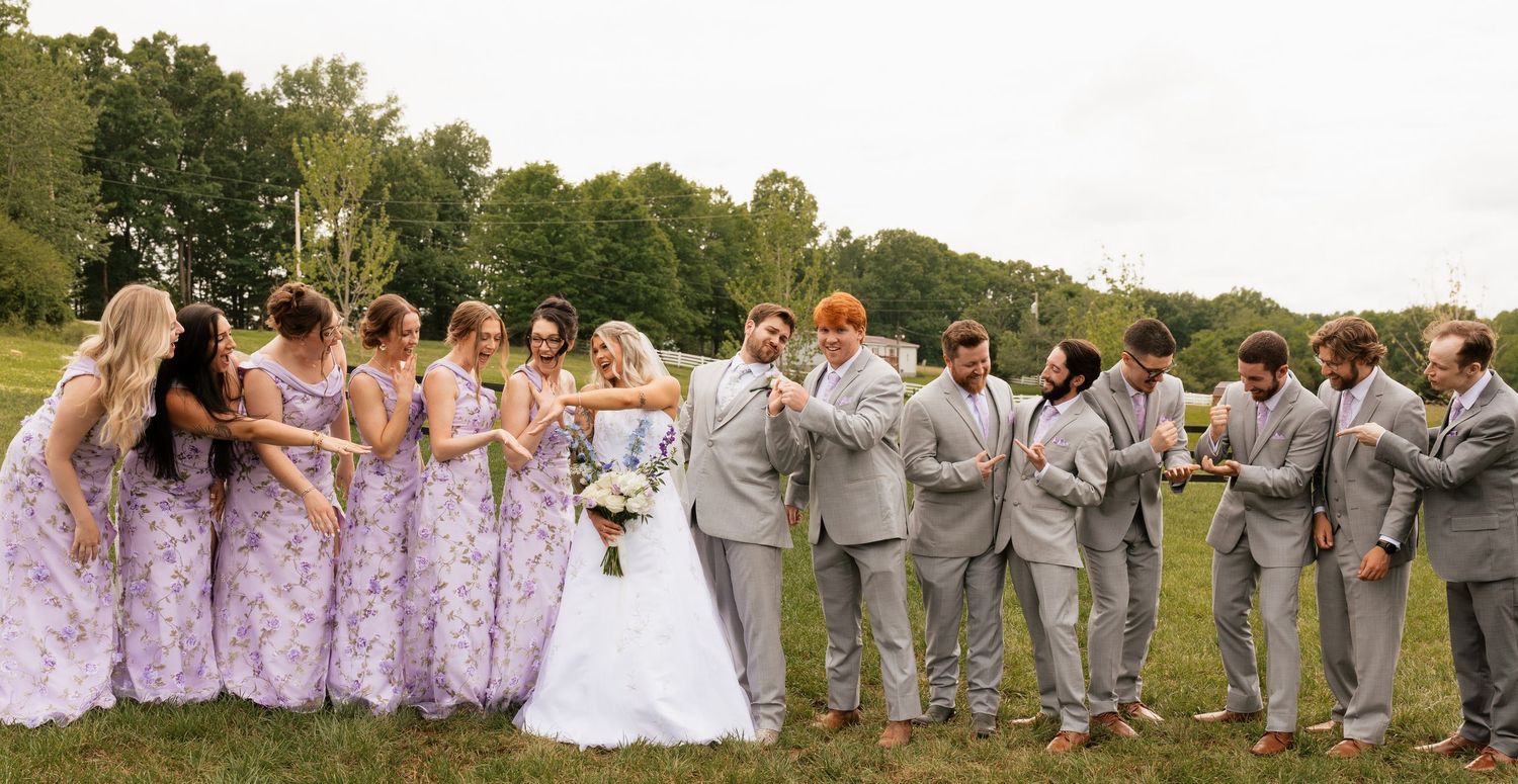 Wedding party poses outdoors, bridesmaids in lavender dresses and groomsmen in gray suits flanking bride and groom.