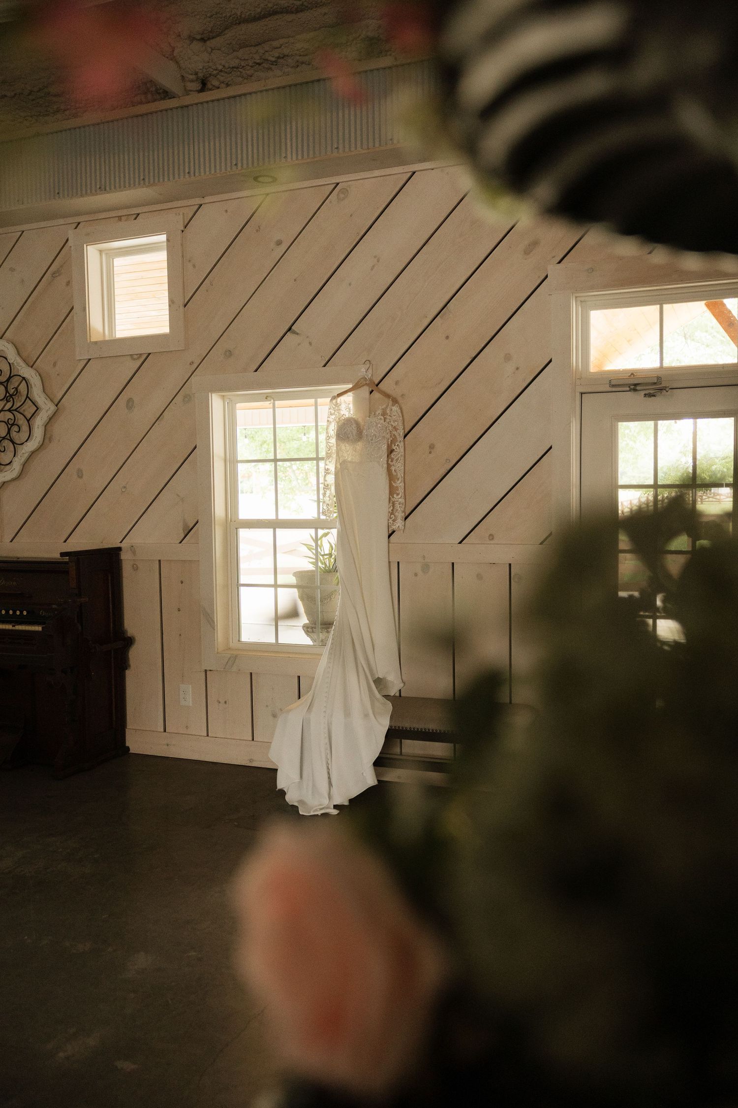 White wedding dress hanging on a rustic whitewashed wood-paneled wall near a window in a charming barn venue.