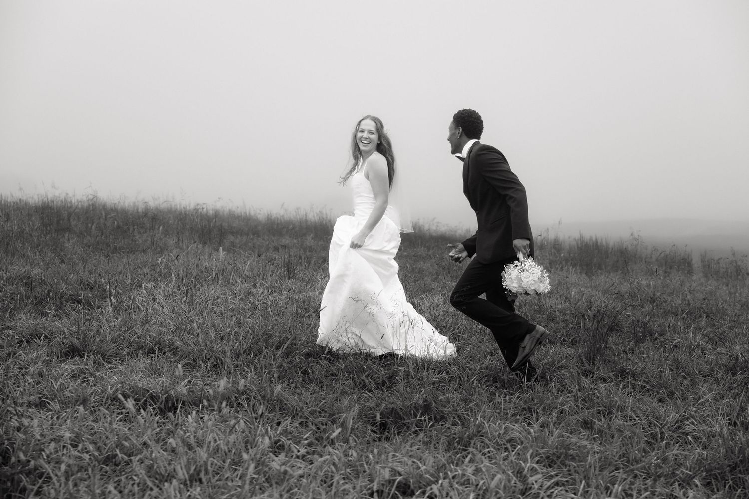 Bride and groom joyfully running through misty field in black and white wedding photo.
