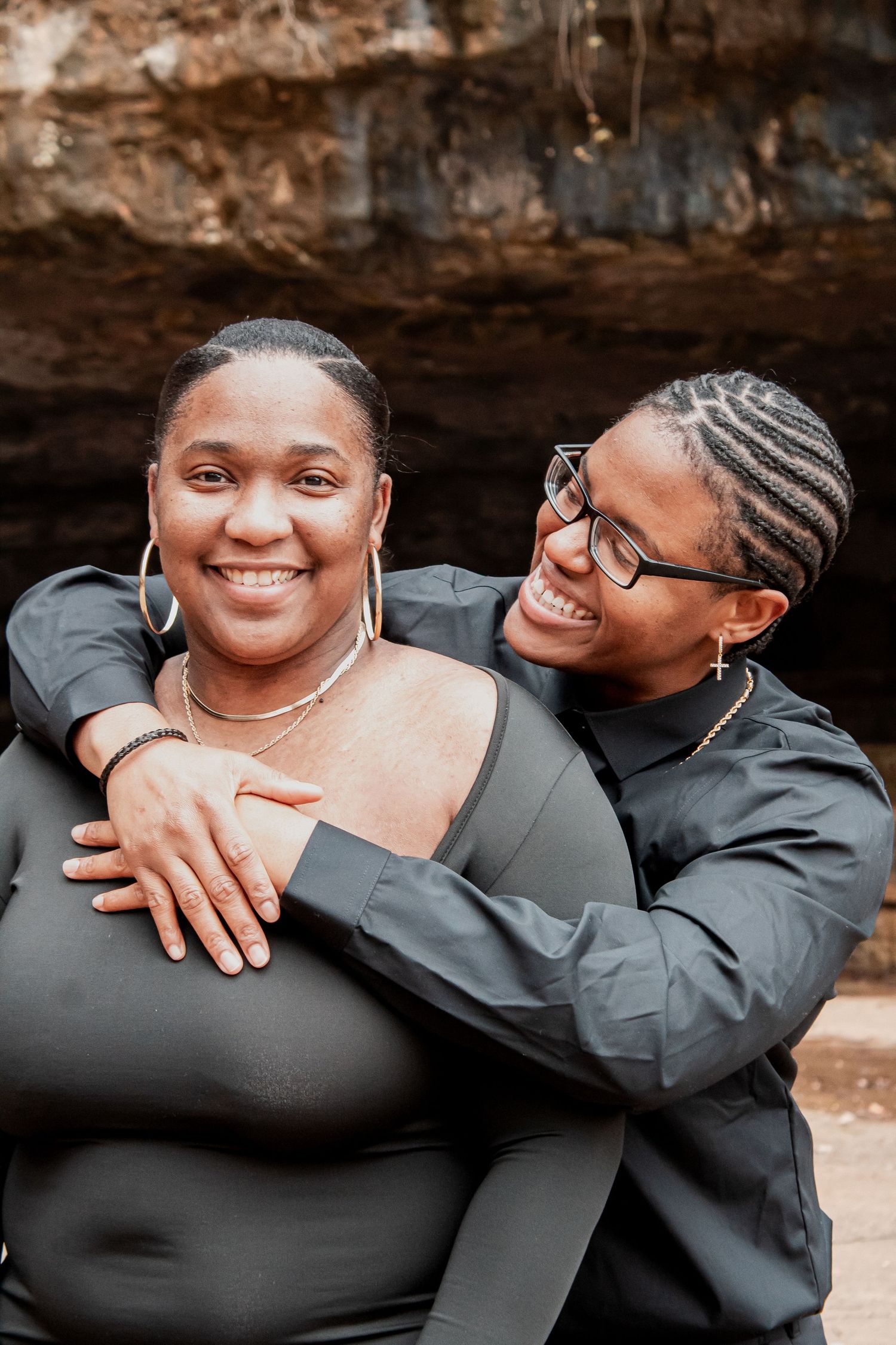 Two smiling women in matching black outfits embracing near a rocky cave backdrop.