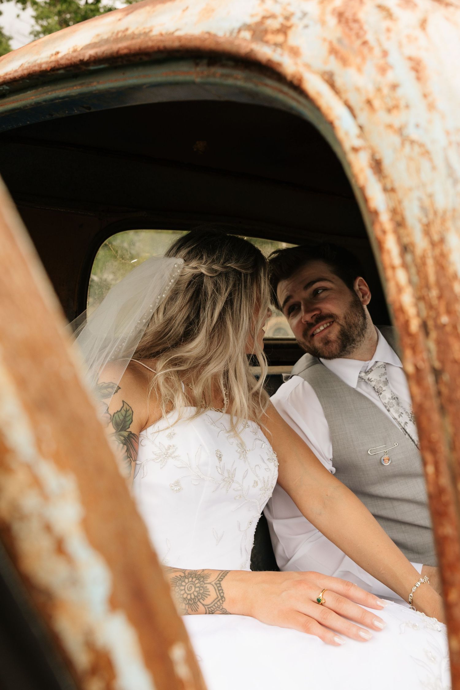 Bride and groom share a romantic moment inside a rustic vintage truck, with the groom smiling at his tattooed bride in her white gown.
