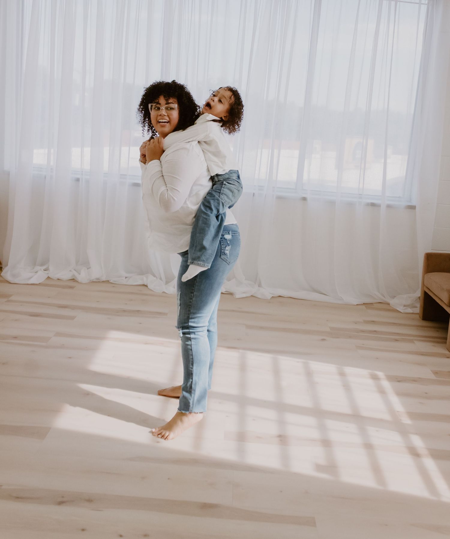 Joyful mother carrying daughter on her back in bright sunlit room with white curtains.