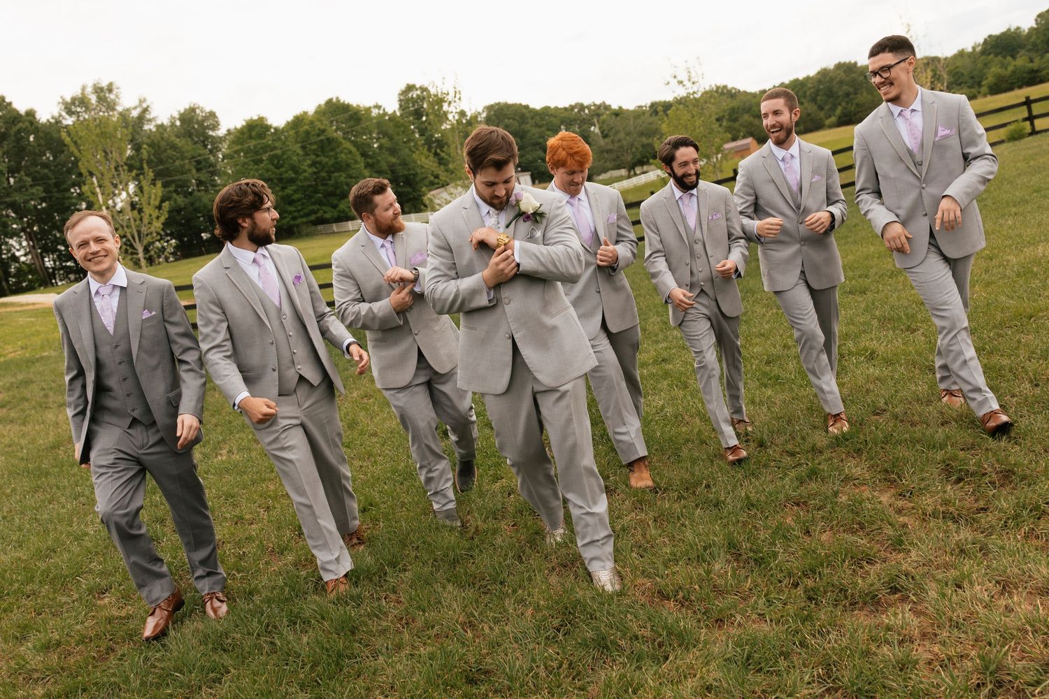 Groomsmen in matching gray suits laughing and posing outdoors on green grass at a wedding celebration.