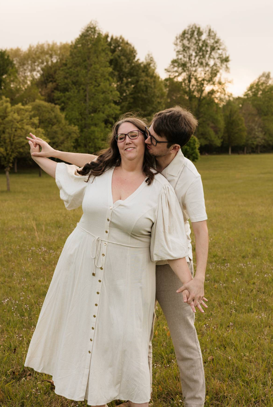 A couple poses in a green field at sunset, woman in white dress pointing while man kisses her cheek.