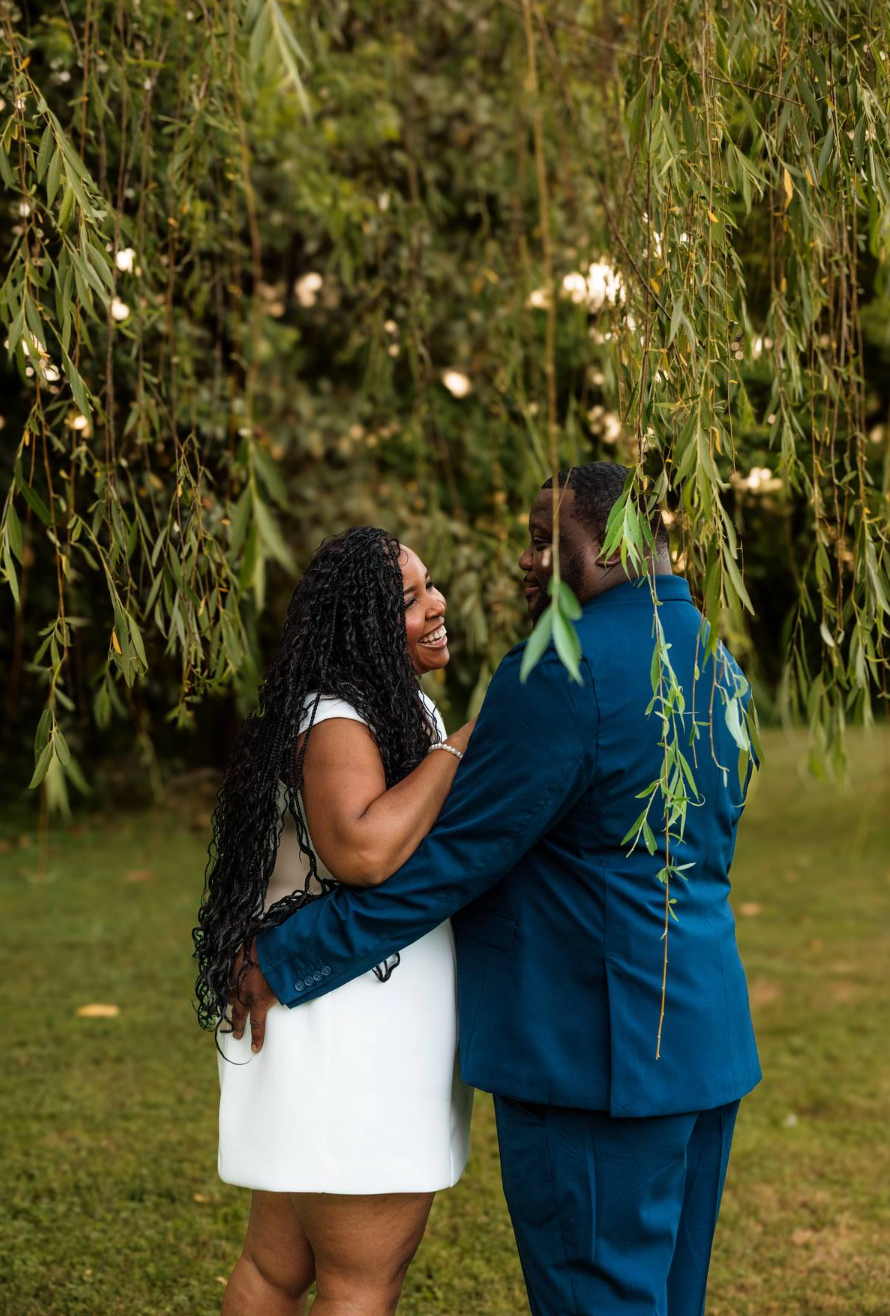 A joyful couple embraces under a weeping willow tree, woman in white dress and man in blue suit.