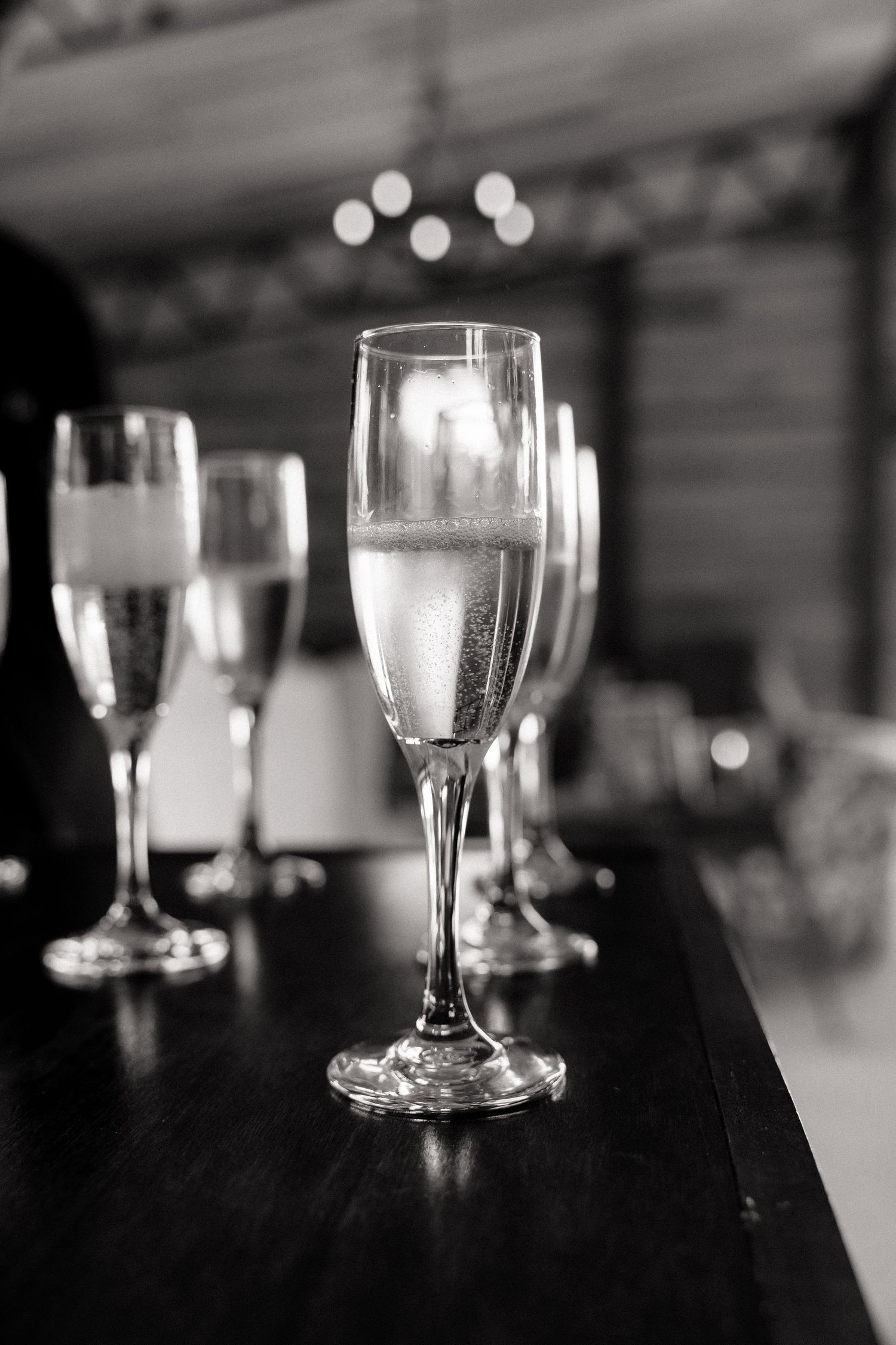 Black and white photo of elegant champagne flutes filled with sparkling wine arranged on a dark wooden bar counter.