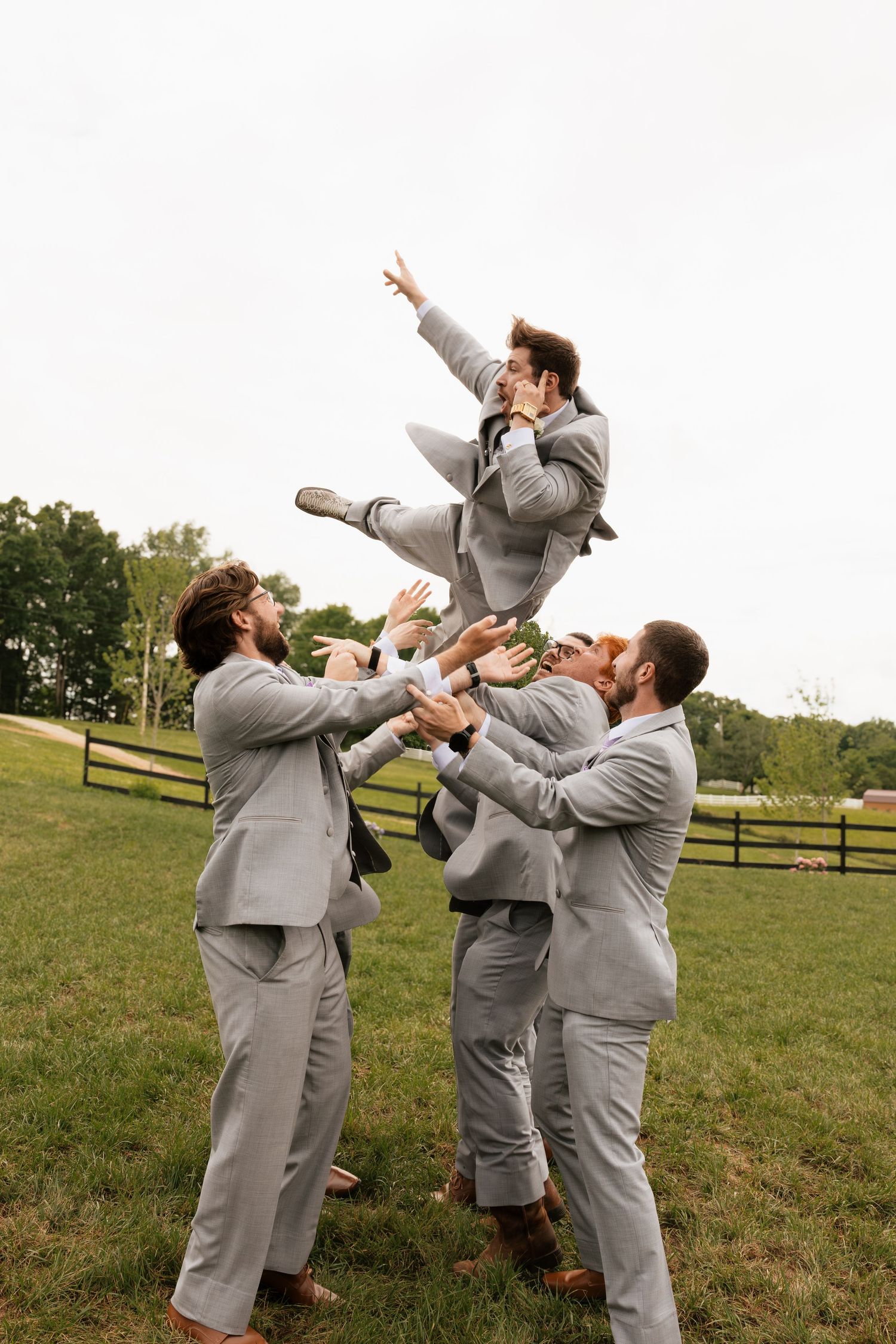 Groomsmen in gray suits tossing the groom into the air outdoors on a green lawn at a wedding celebration.