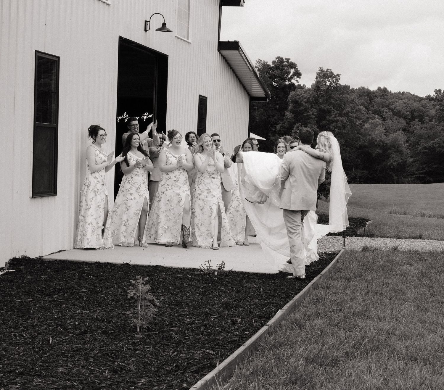Black and white photo of bride with bridesmaids in floral gowns outside a white barn venue.