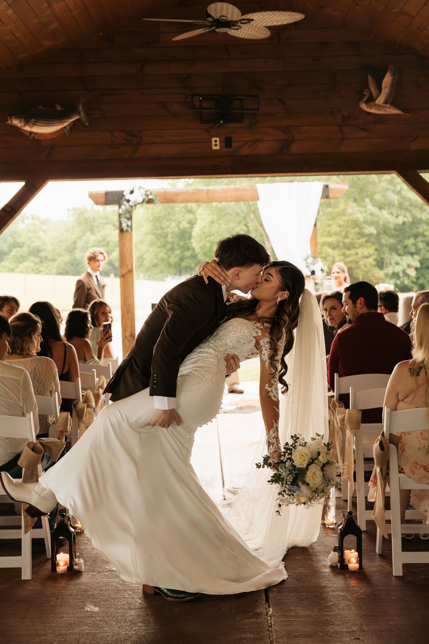 Bride and groom share a romantic dip kiss at the altar during their rustic barn wedding ceremony with guests seated nearby.
