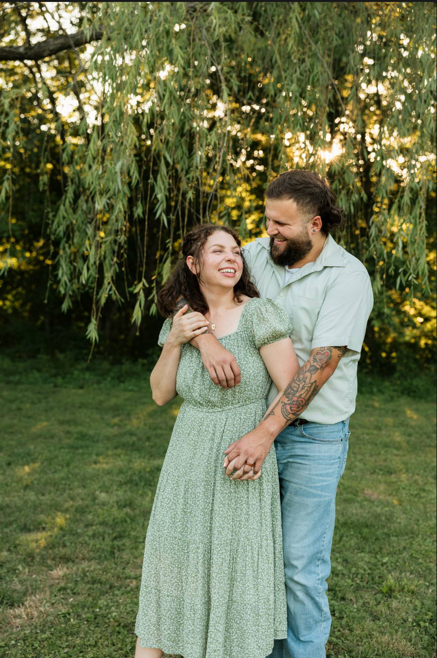 A smiling couple embraces outdoors near a weeping willow tree, woman in green floral dress, man with tattoos in light blue shirt.