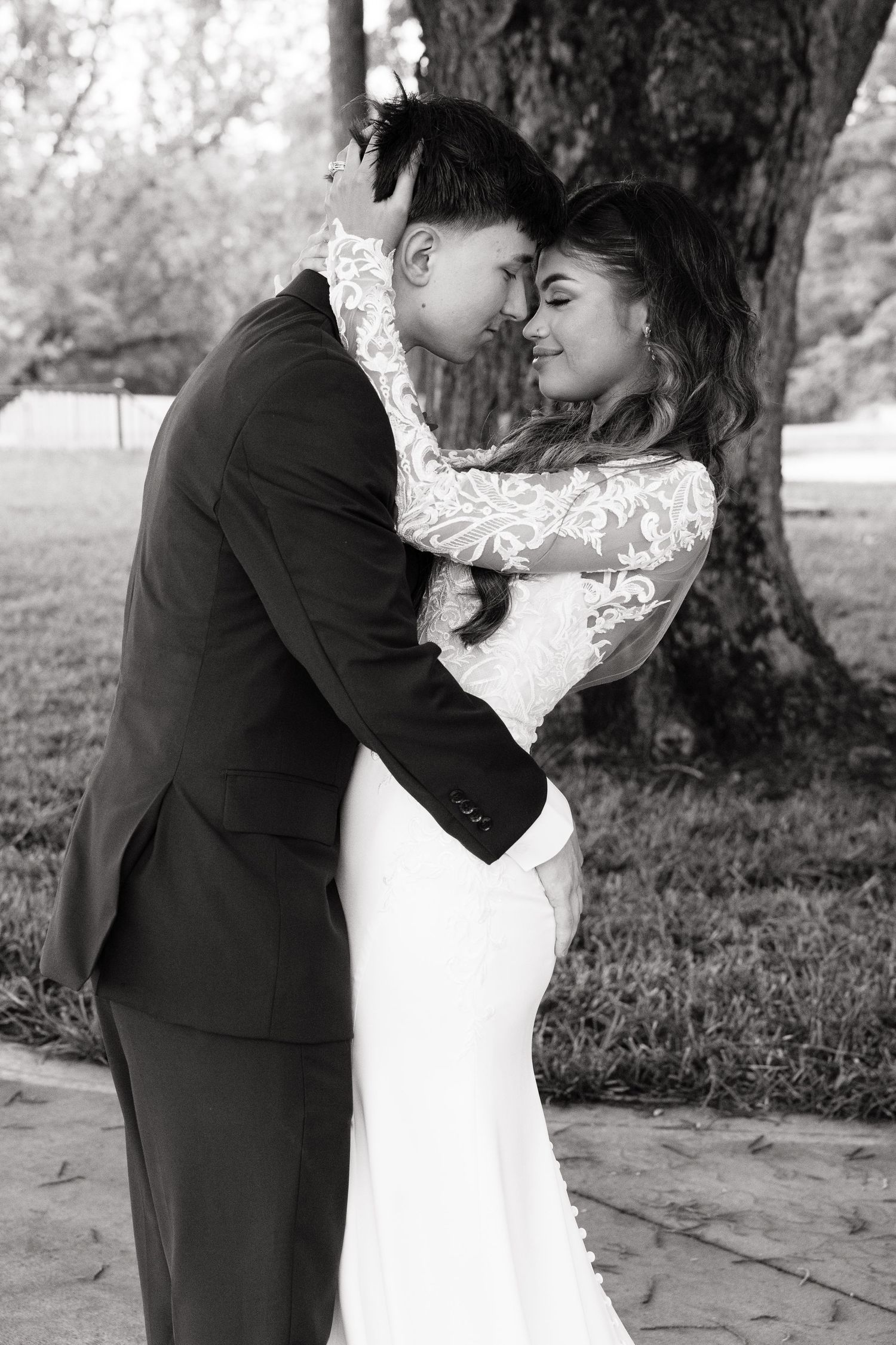Black and white photo of a bride and groom embracing tenderly outdoors near a large tree on their wedding day.