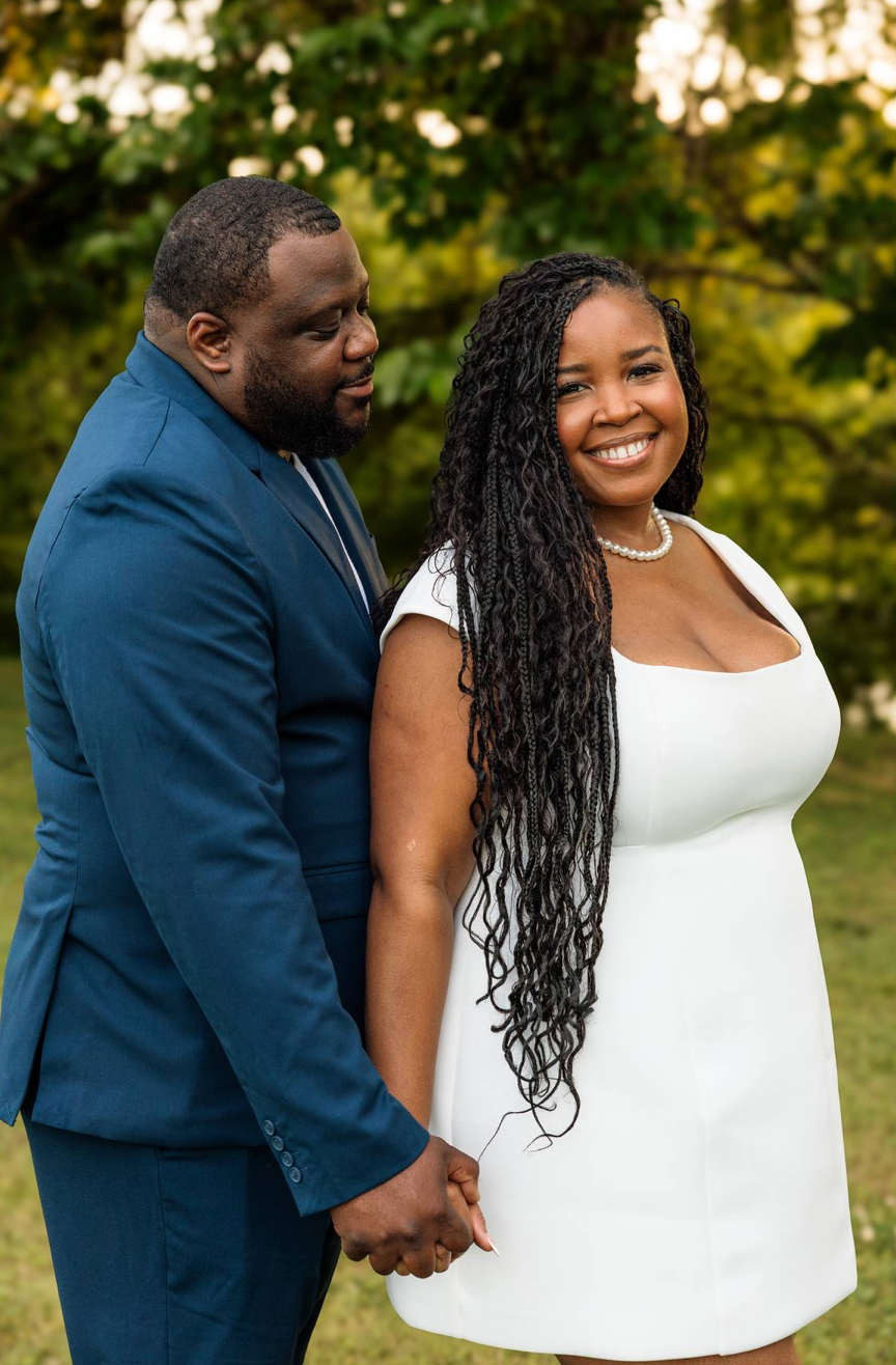 A smiling couple holds hands outdoors, woman in white dress with long braids, man in blue suit, green trees behind.