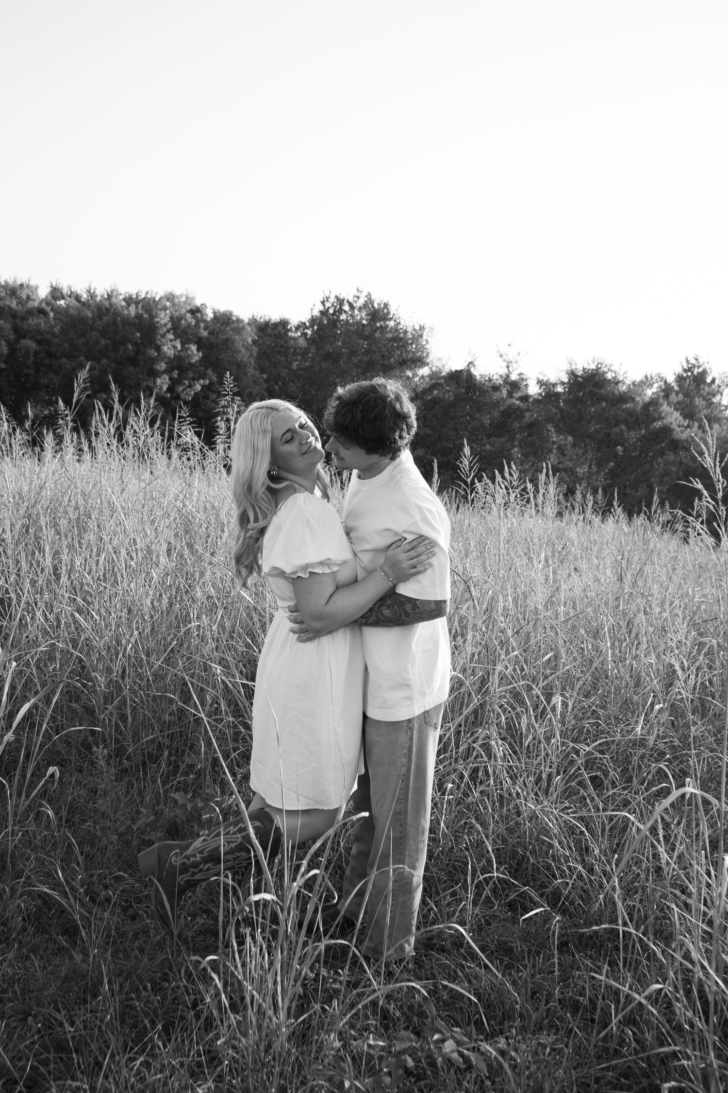 Black and white photo of a couple embracing romantically in a tall grass field with trees in the background.