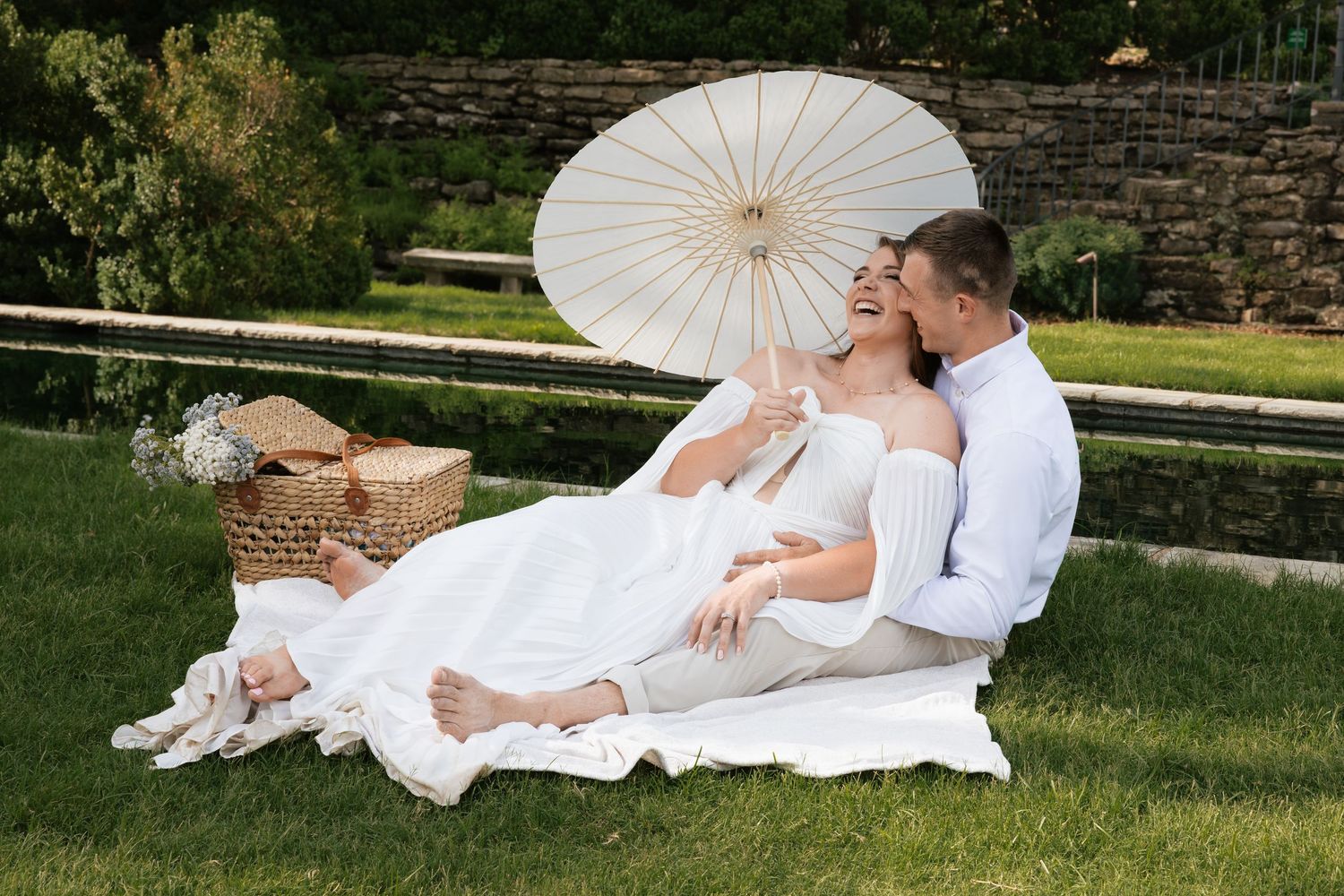 Newlywed couple in white attire enjoying a romantic outdoor picnic on grass with a parasol umbrella.